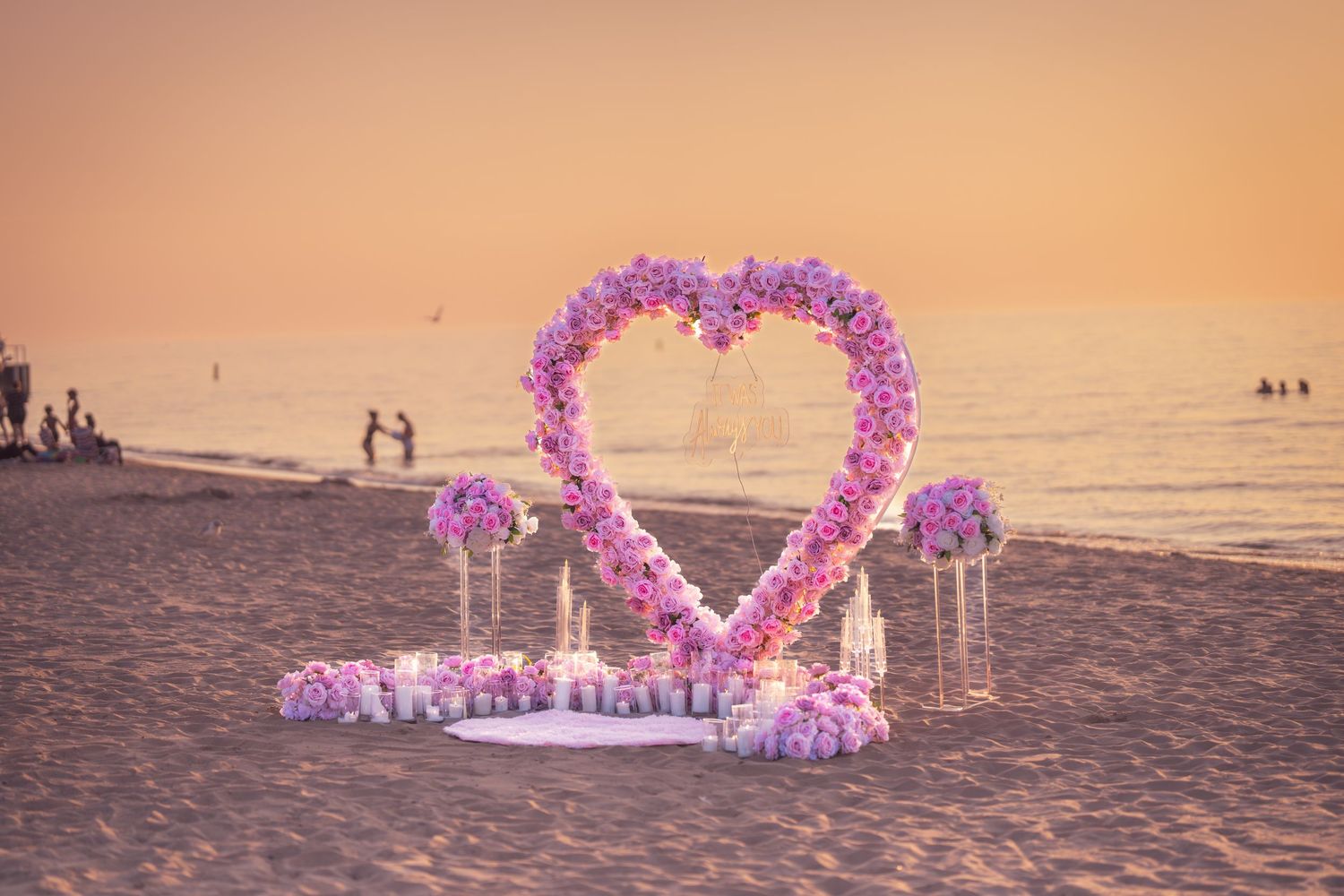 A romantic beach proposal setup with pink balloon arch and decorations during sunset by the ocean.