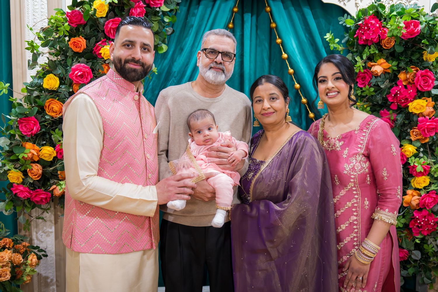 South Asian family of five with baby posing at floral arch during festive occasion.