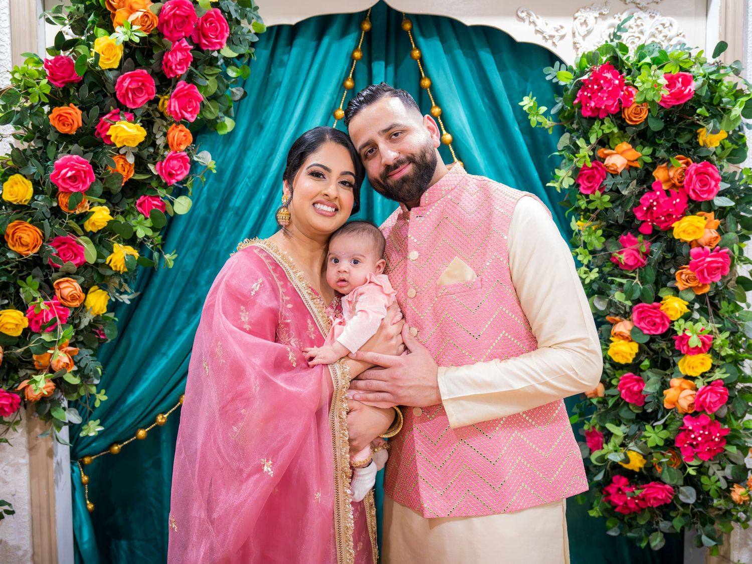 Indian family in traditional attire poses with baby before colorful floral arch with teal drape backdrop.
