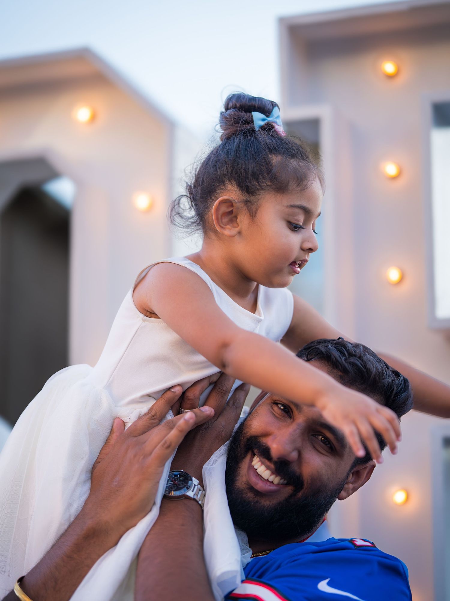 A bearded man in a blue Bills jersey joyfully lifts a smiling toddler girl onto his shoulders outdoors.