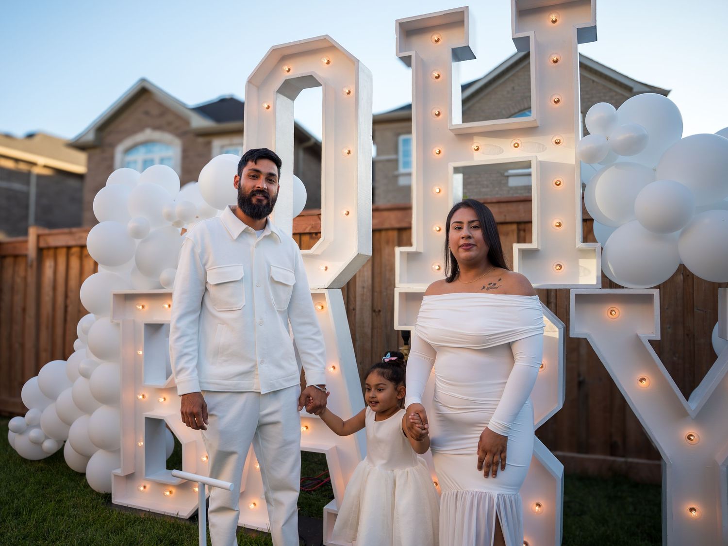 Family of three in white outfits posing in front of illuminated OH BABY marquee letters with balloon arch at gender reveal party.