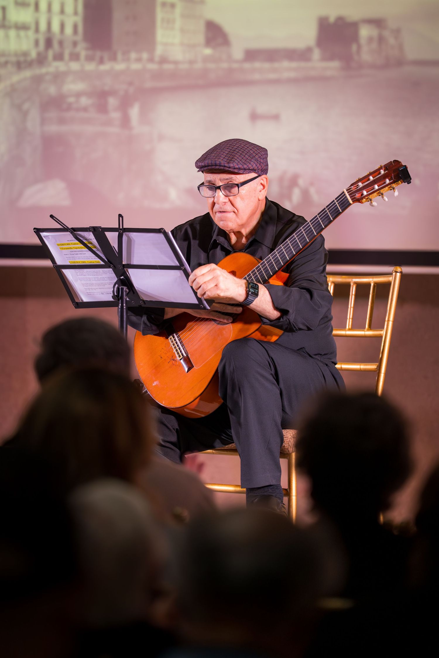 An elderly guitarist in a cap and glasses plays classical guitar on stage for a seated audience.