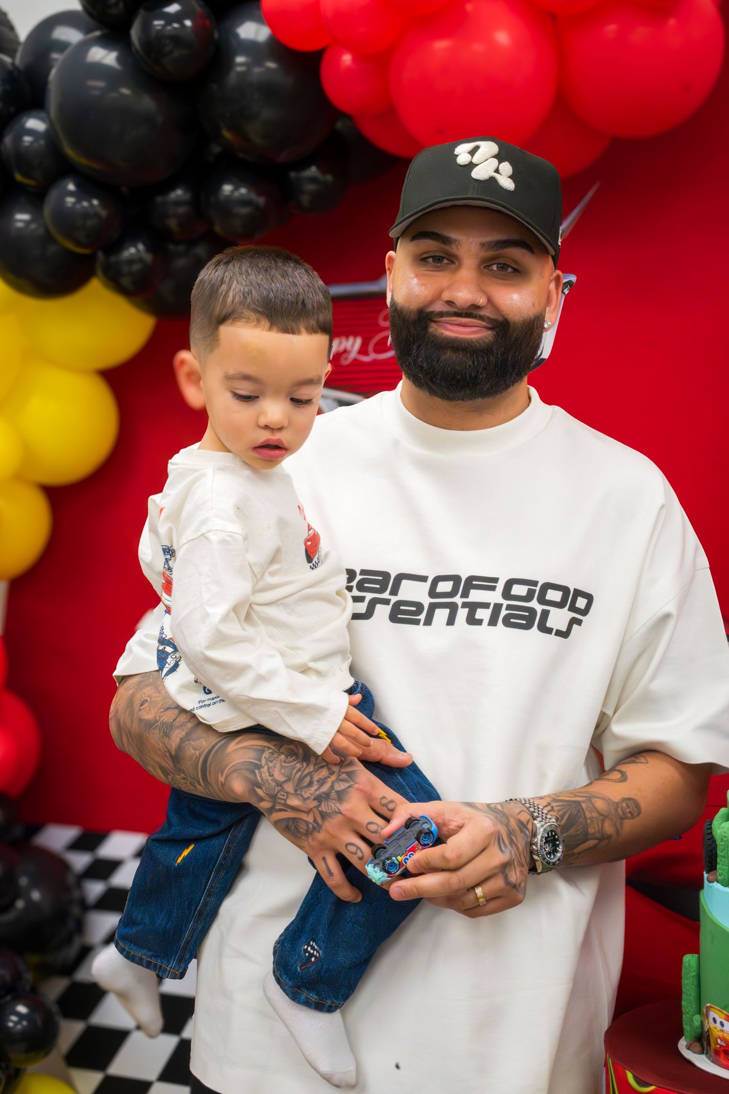 A tattooed man in a Fear of God Essentials tee holds a toddler at a Cars-themed birthday party.