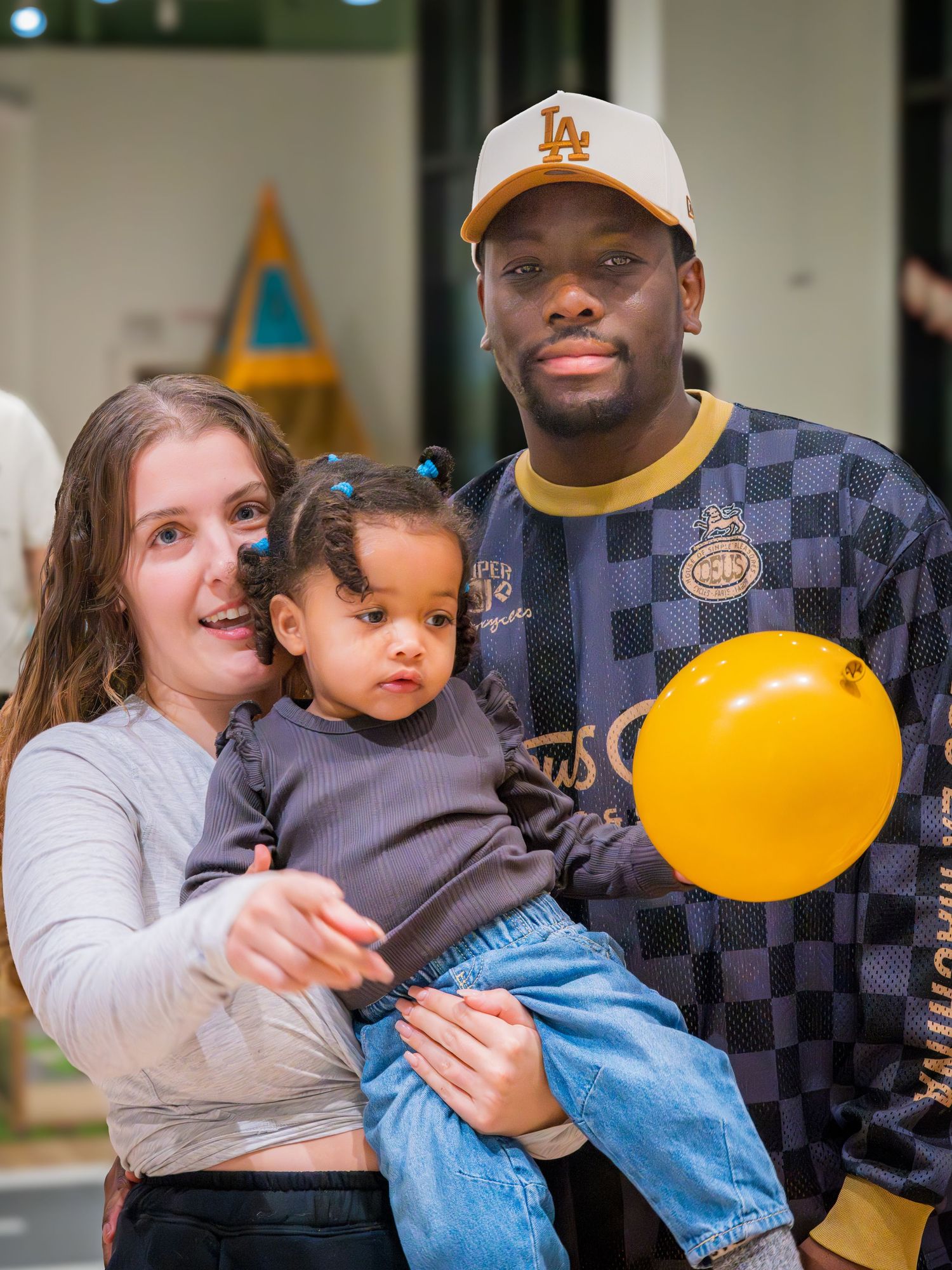 Family of three with parents holding their baby girl with a yellow balloon at an indoor children's event.