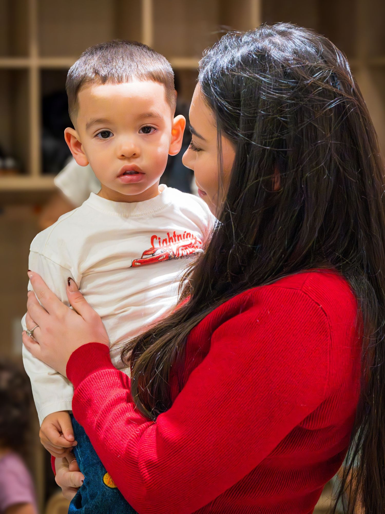 A smiling dark-haired woman in a red sweater lovingly holds a toddler boy in a Lightning McQueen shirt.