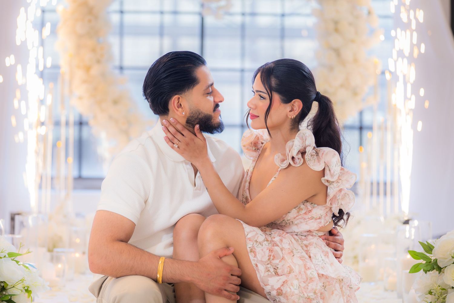 Couple sharing a tender moment seated together beneath a glowing white floral arch.