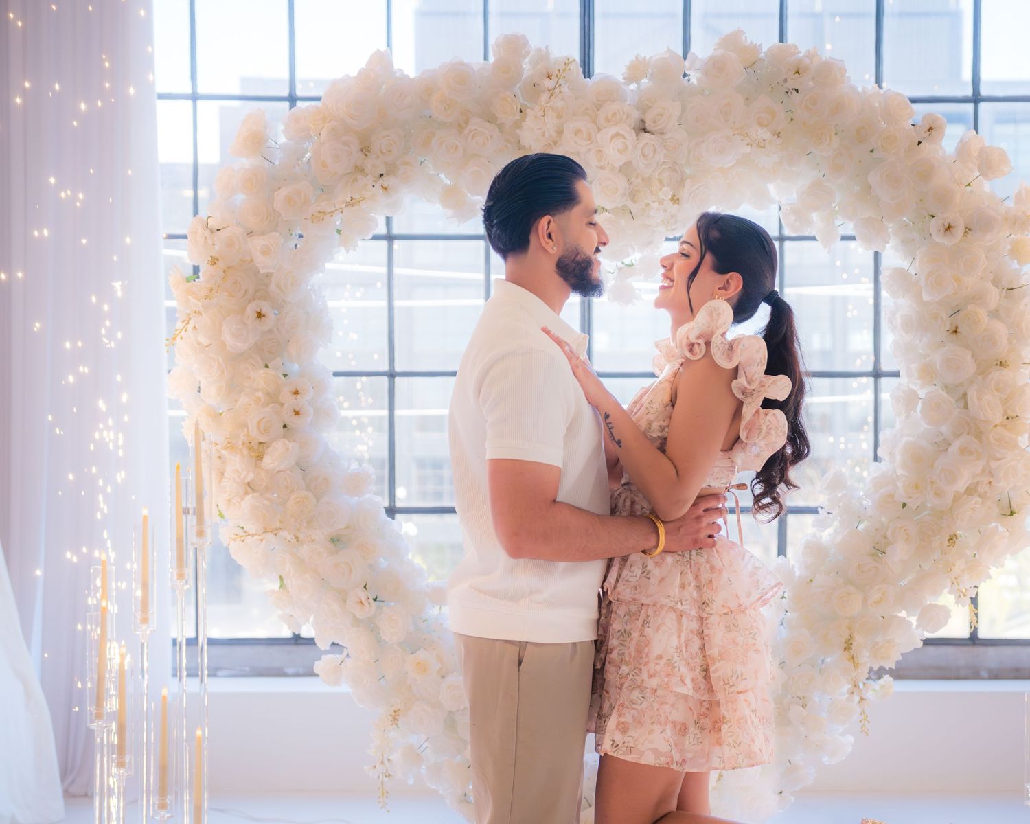 Couple gazing into each others eyes framed by a white rose heart arch with fairy lights.