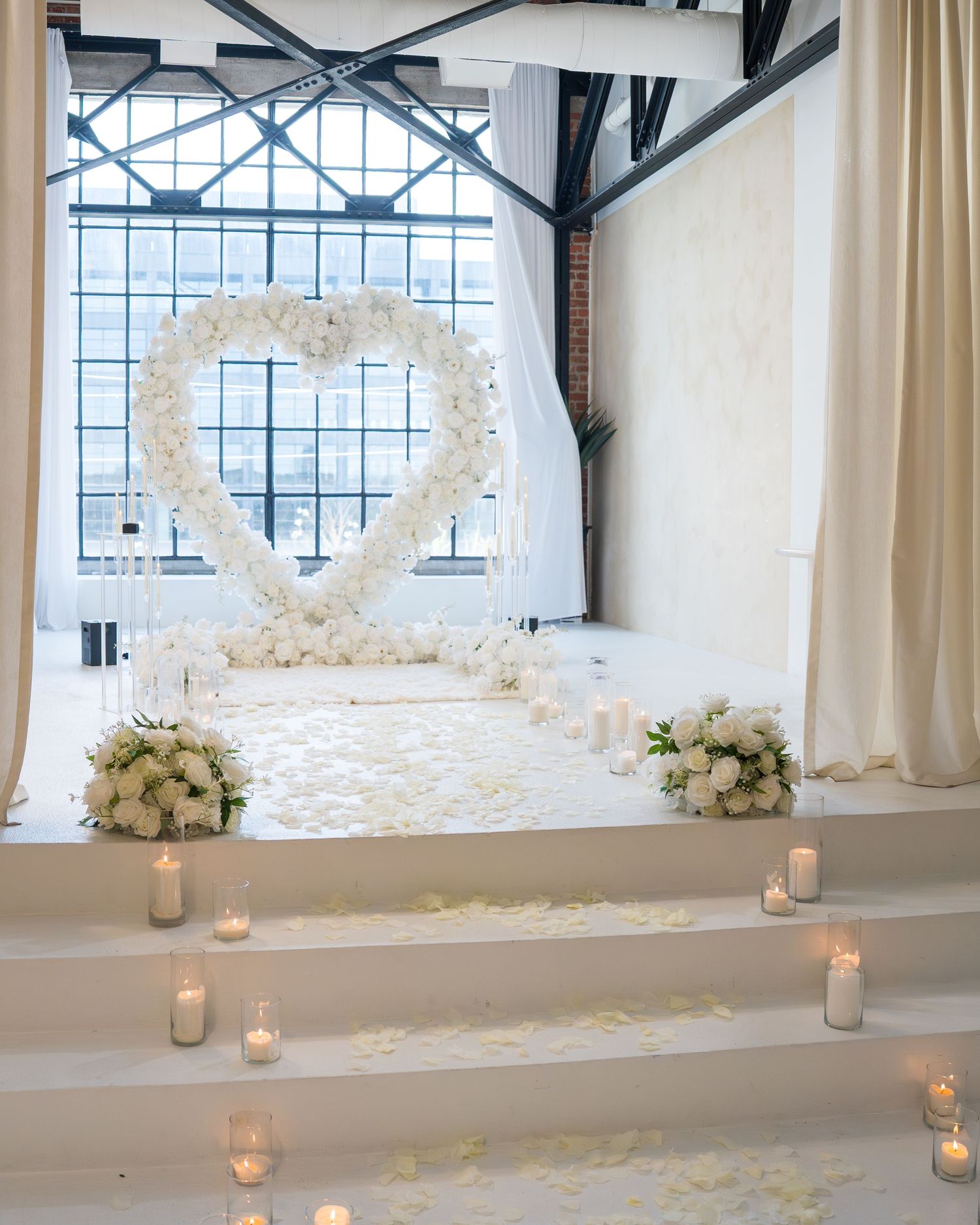 White floral heart arch wedding ceremony backdrop with candles and rose petals on white steps.
