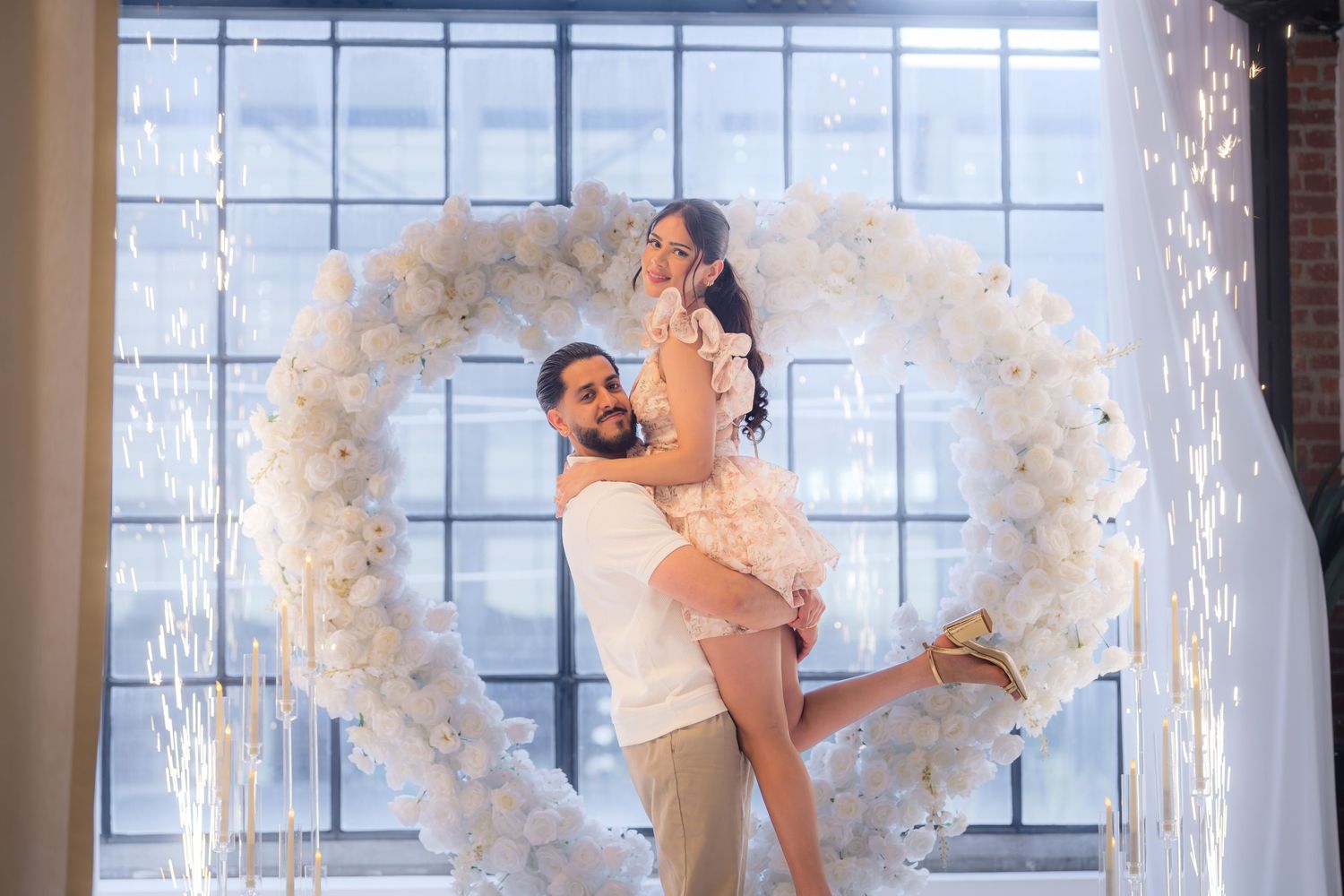 Man lifting smiling woman inside a heart-shaped white floral arch with celebration sparklers.