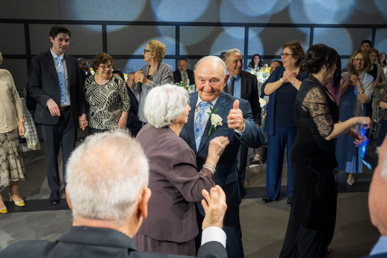 Elderly couple dancing joyfully at a formal event surrounded by smiling guests in a modern venue.