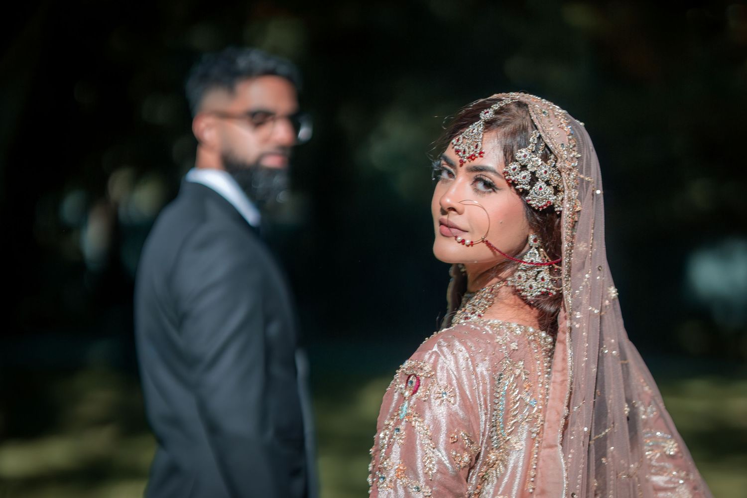 Artistic portrait of bride in embellished dress with groom blurred in background.