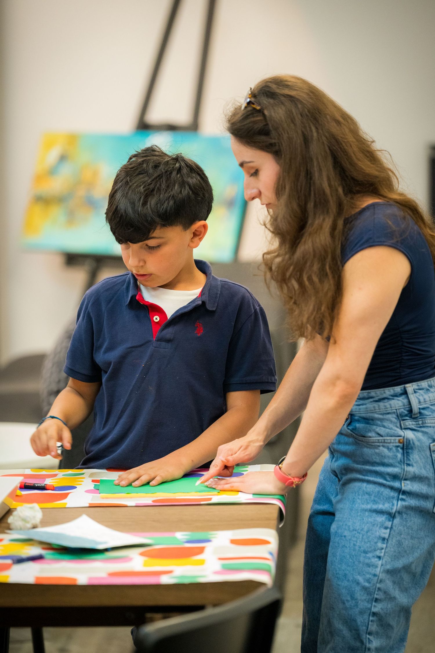A woman helps a young boy with a colorful paper craft project at a table in an art classroom.