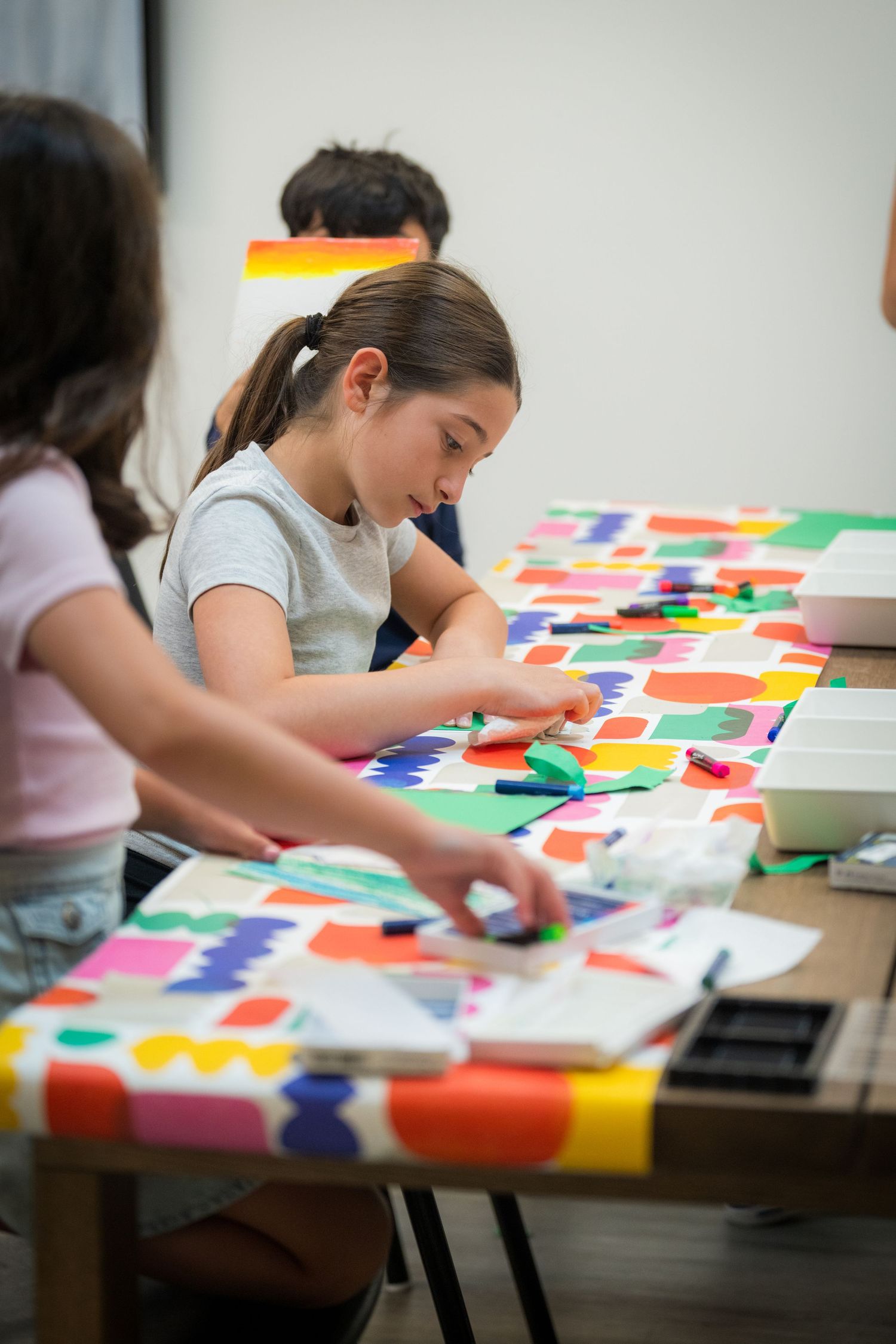 Girl focuses on colorful paper craft project at art table covered with bright shapes.