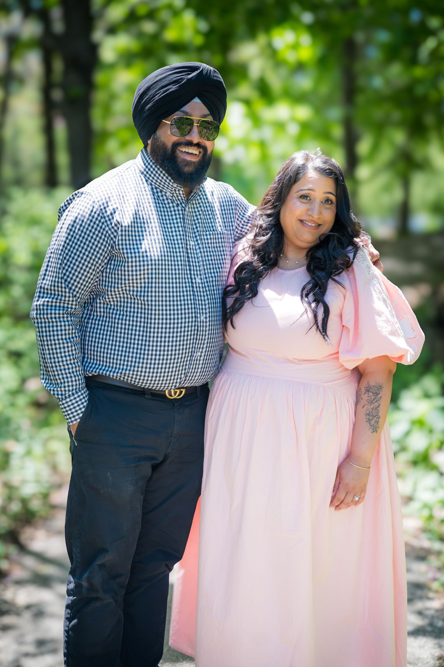 A smiling Sikh man with sunglasses and a woman in a pink dress pose together in a park.