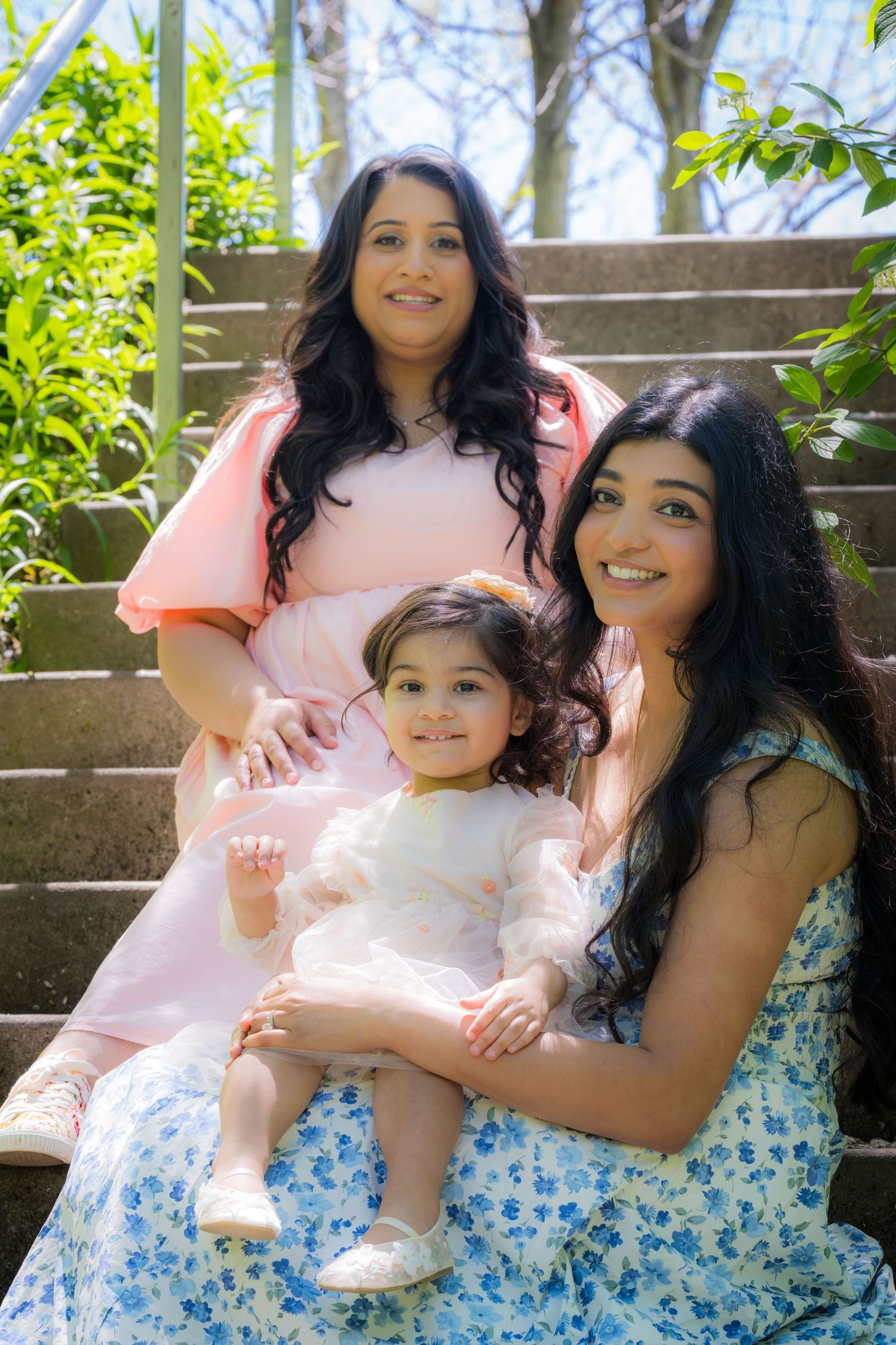 Two women and a toddler in white dress pose together on outdoor stone steps in park.