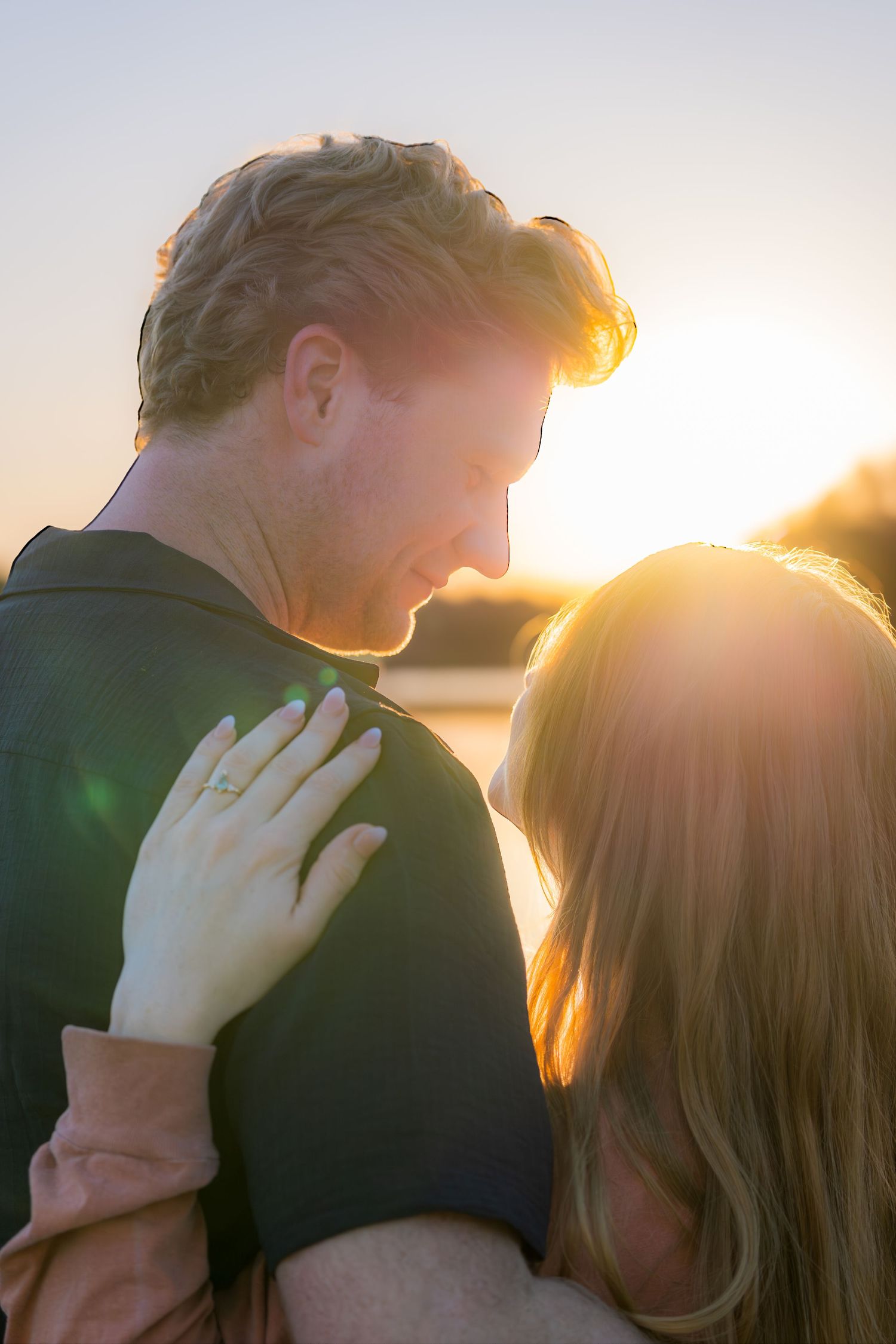 Couple embracing at sunset with golden light and lens flare, womans hand showing engagement ring.