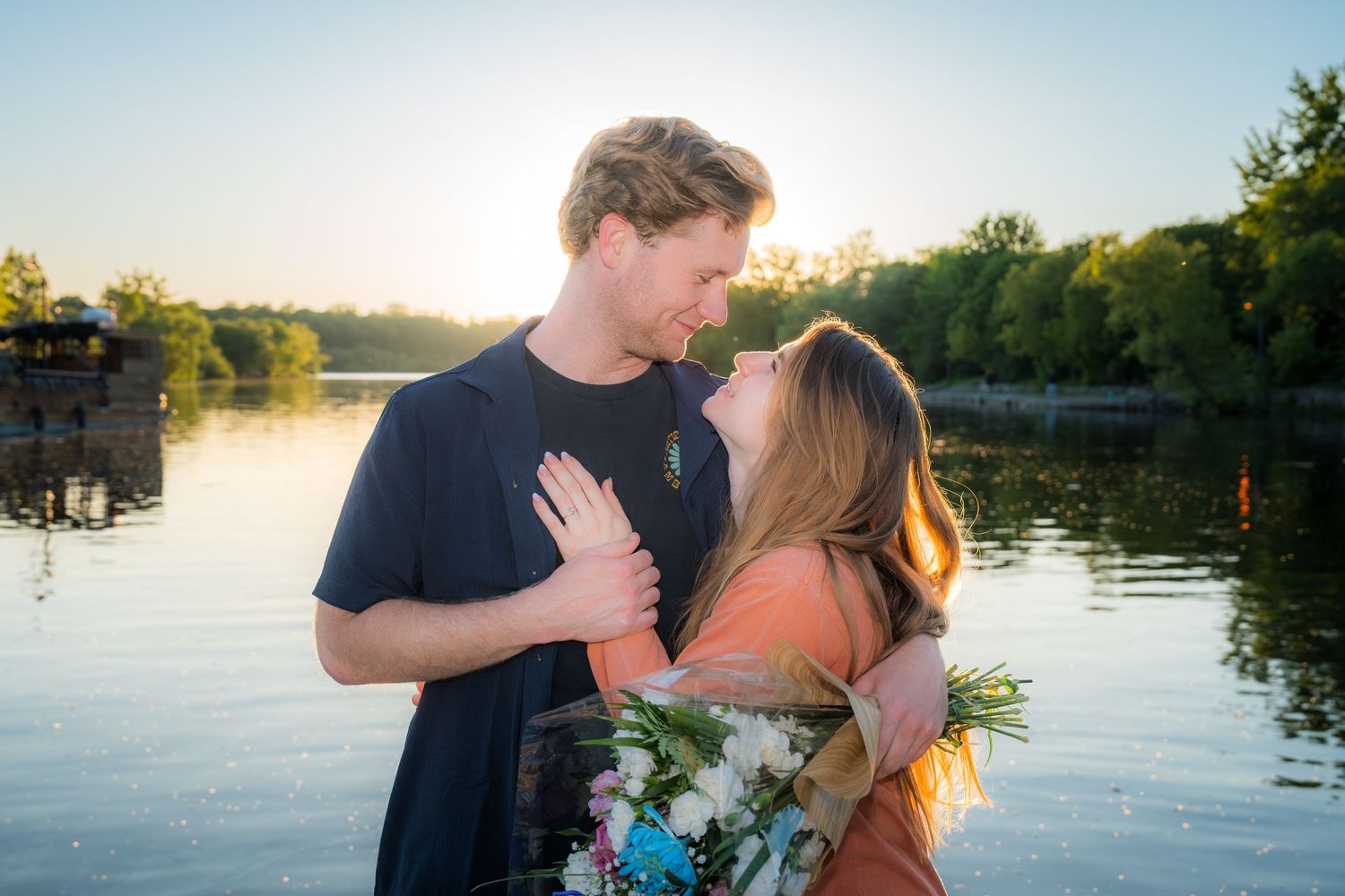 Couple sharing a romantic moment near a sunlit lake, man holding woman with a bouquet of flowers at golden hour.