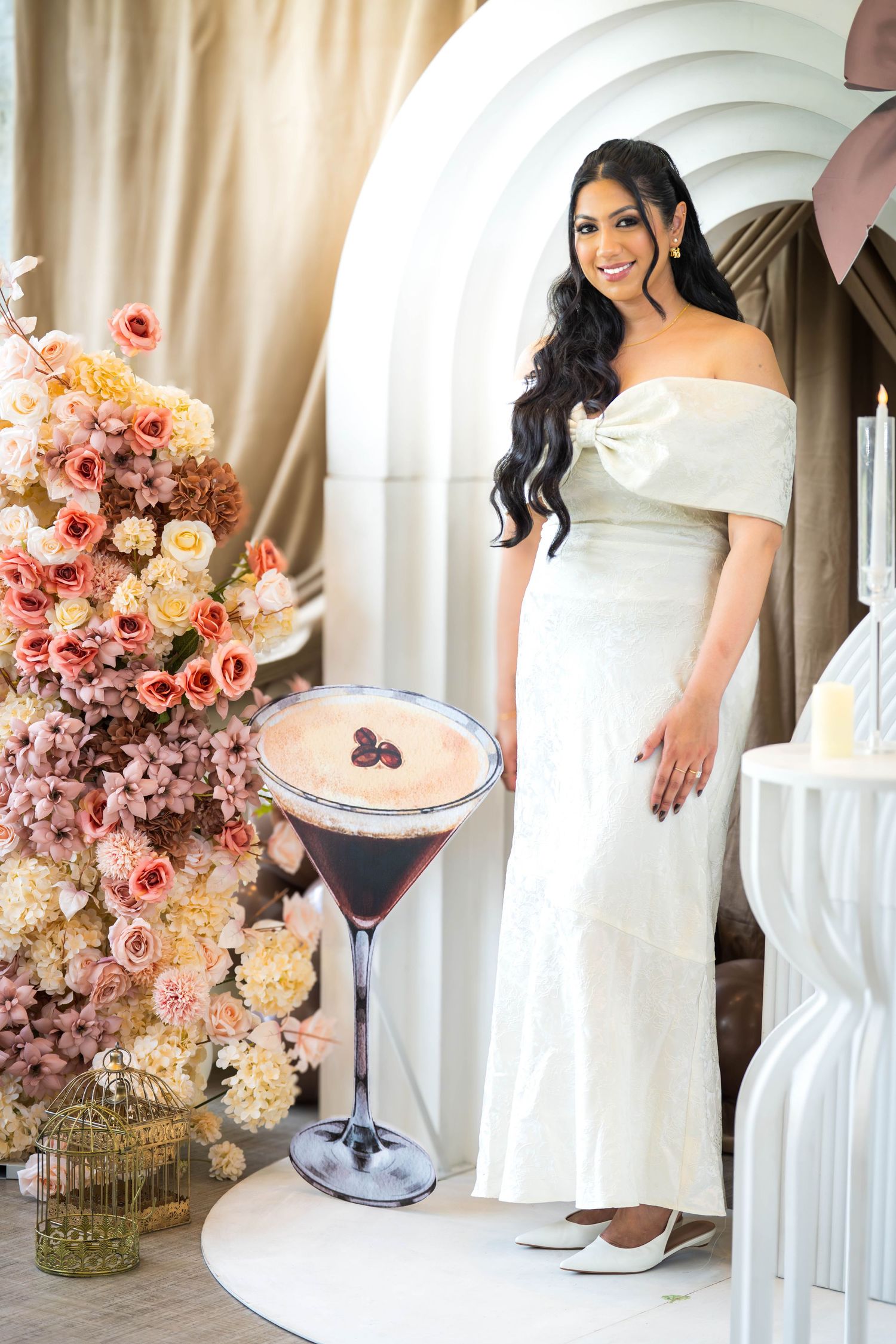 Woman in white off-shoulder gown poses beside oversized espresso martini and floral arrangement at elegant event.