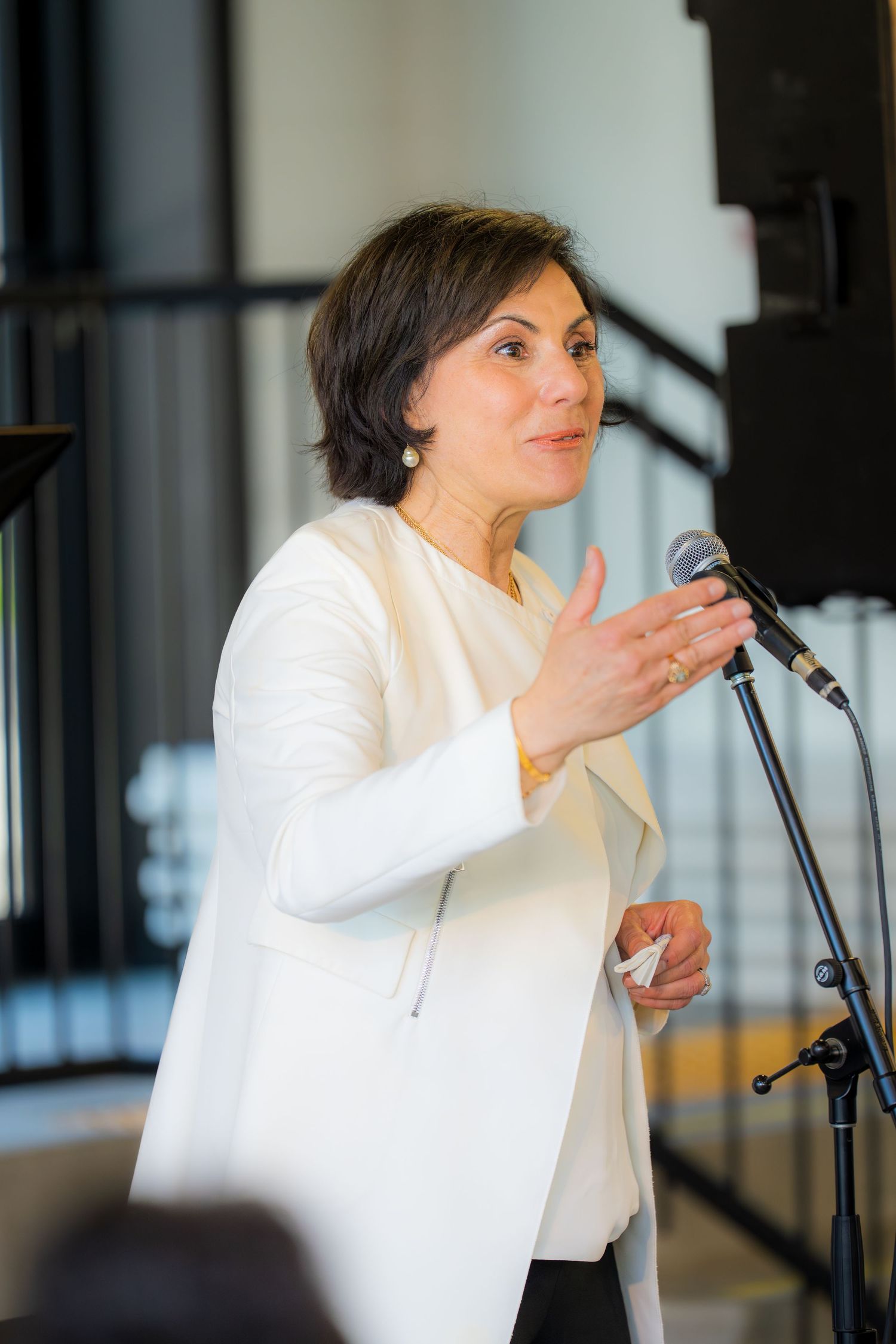 Woman in white blazer speaking at microphone during indoor event, gesturing with hand.