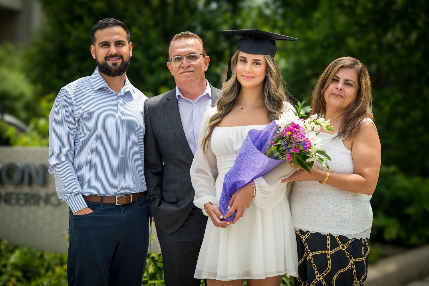 A graduate in white cap and gown holds a purple bouquet while posing outdoors with family members.