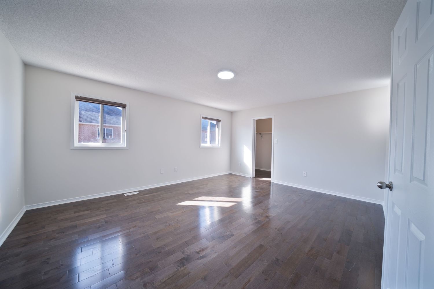 Empty room with hardwood floors, white walls, and two small windows showing natural light streaming in.