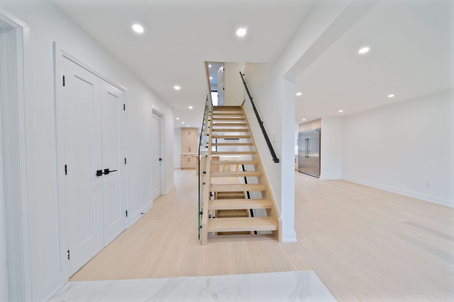 Modern open staircase with wooden treads and glass railings in a bright white interior hallway.