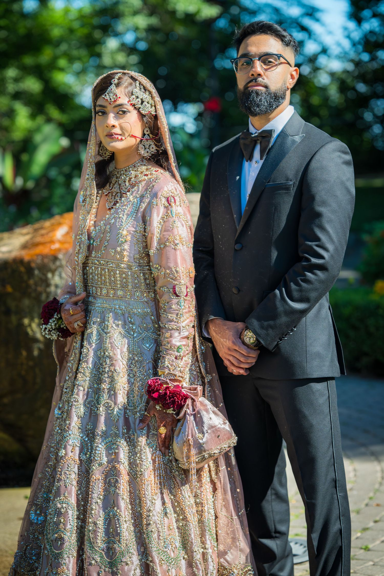 Bride in ornate peach lehenga and groom in black suit stand together in garden setting.