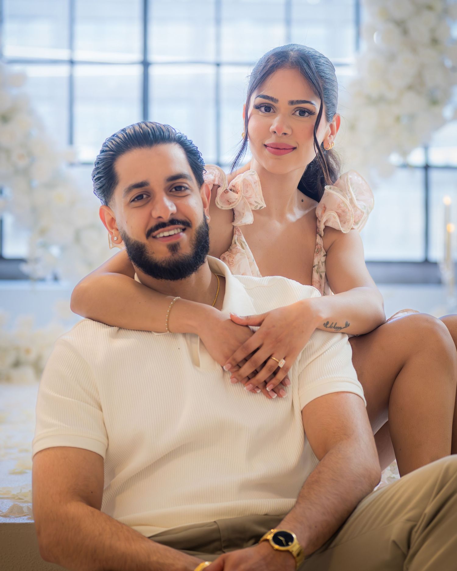 Engaged couple posing together with white floral decorations in the background.