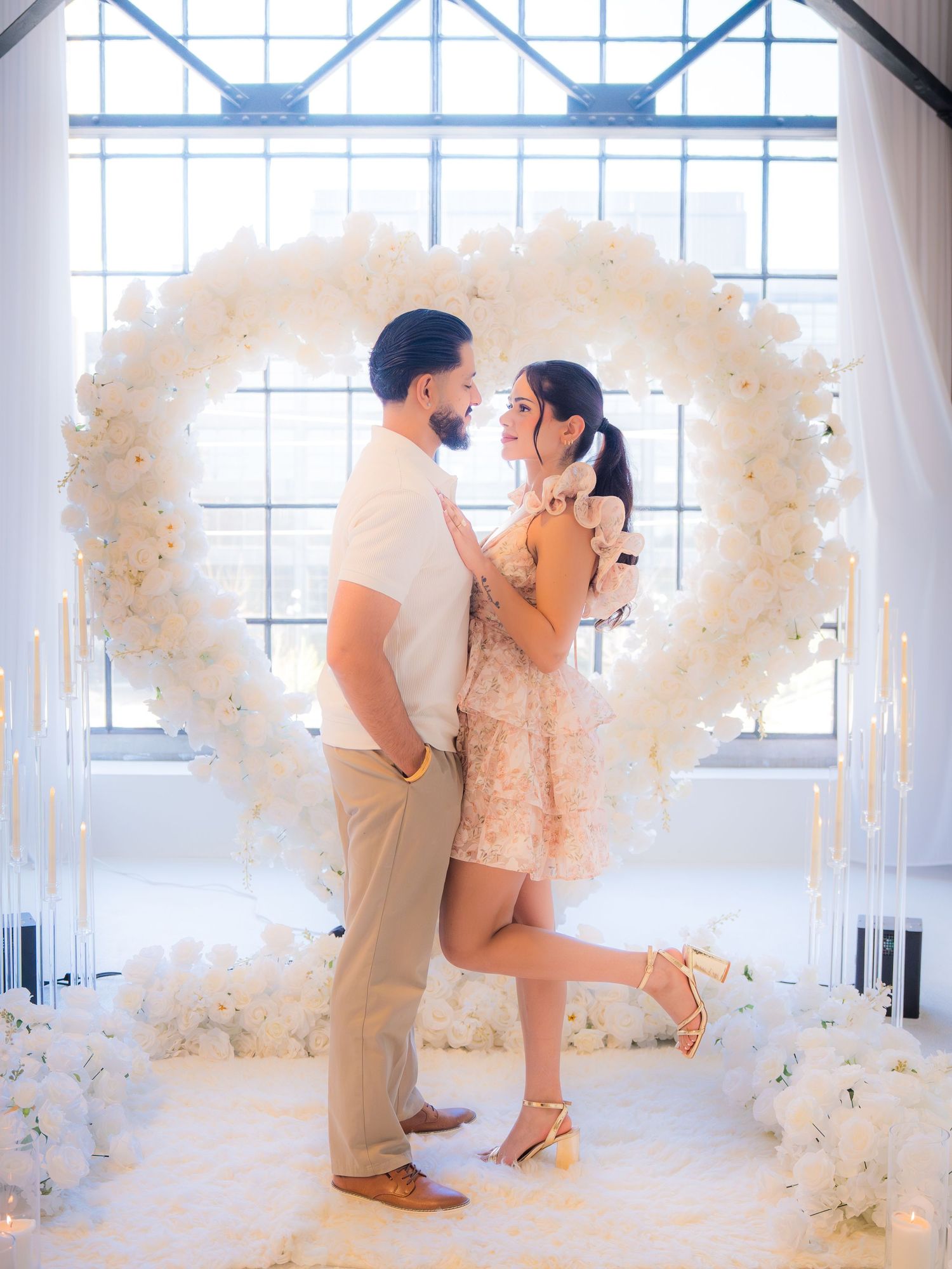 Couple embracing and smiling in front of large white floral heart arch with candles and flowers.