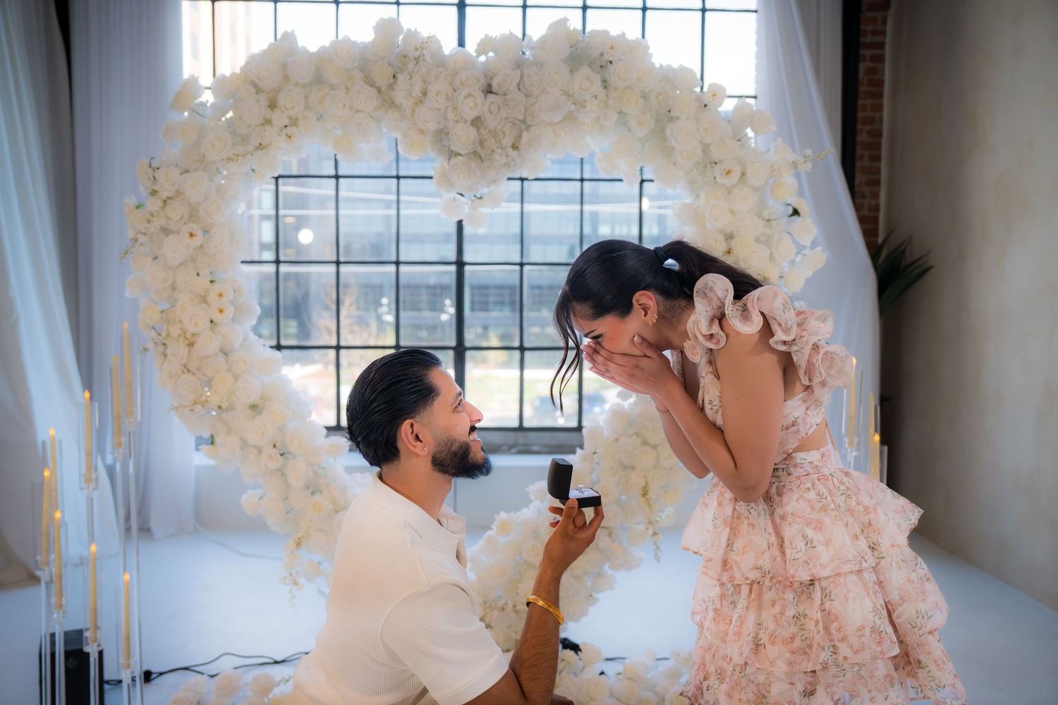 Man proposing to woman in front of a stunning white floral heart arch with candles.