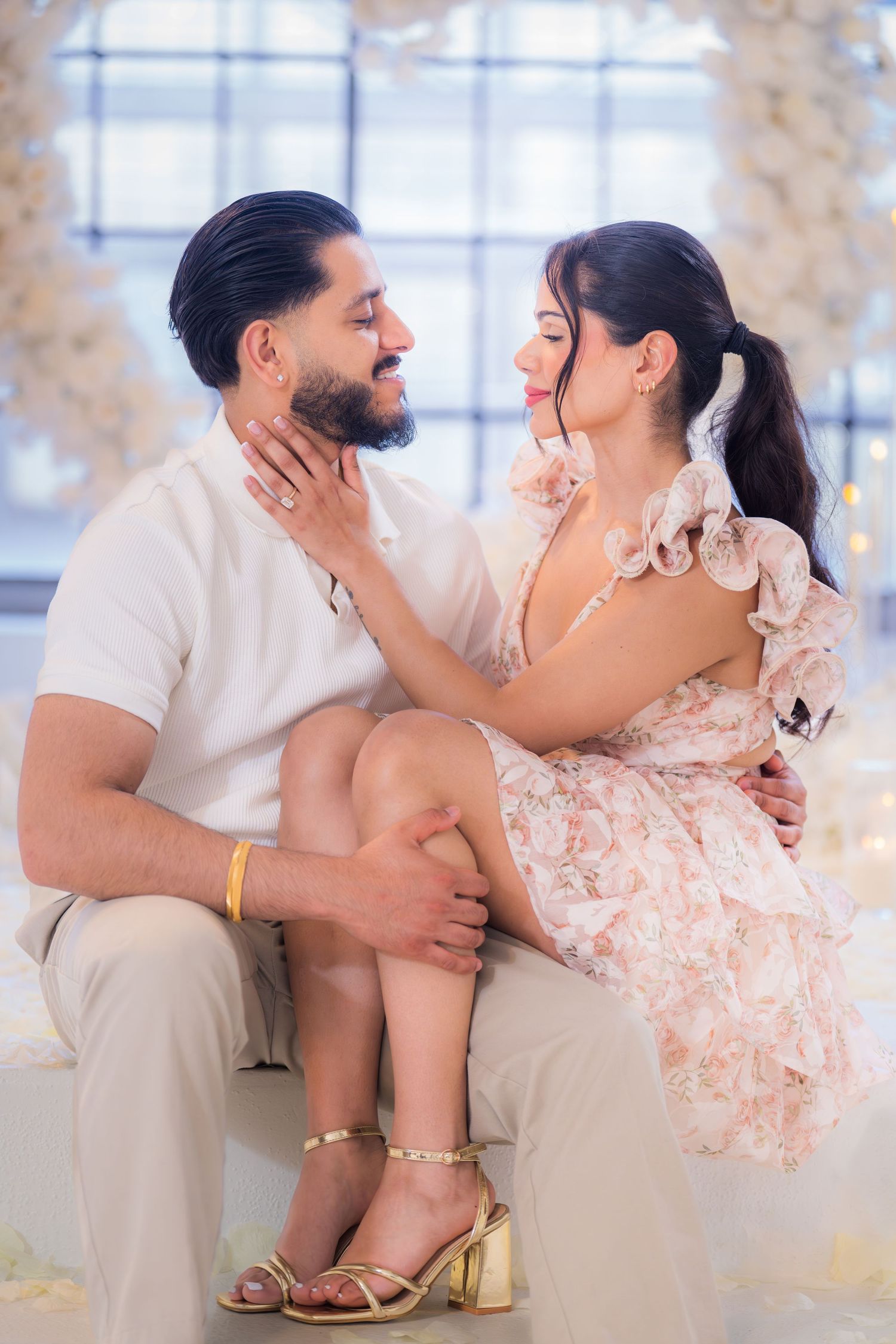 Woman sitting on mans lap tenderly touching his face in romantic floral setting.