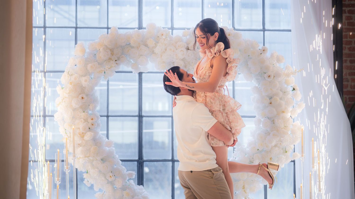 Man lifting woman in floral dress inside a white balloon heart arch with sparkling effects.
