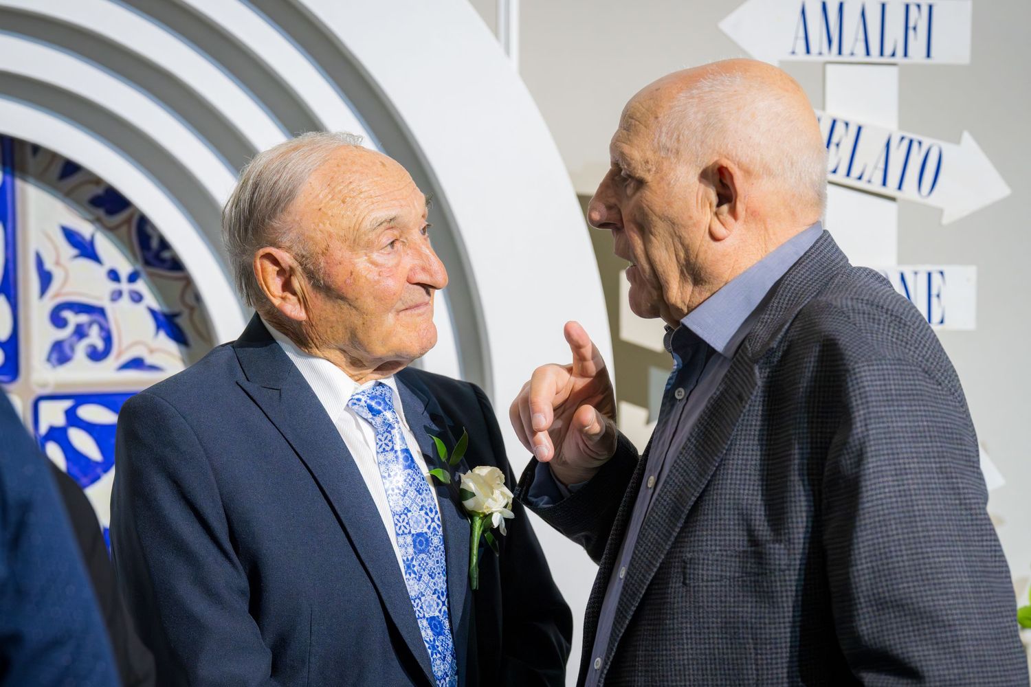 Two elderly men in suits having a conversation outdoors near decorative Italian-themed signage.