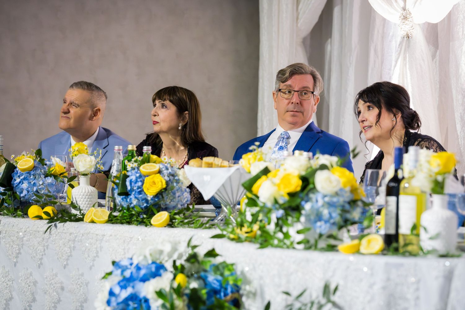 Four guests seated at a wedding head table adorned with yellow and blue floral centerpieces.