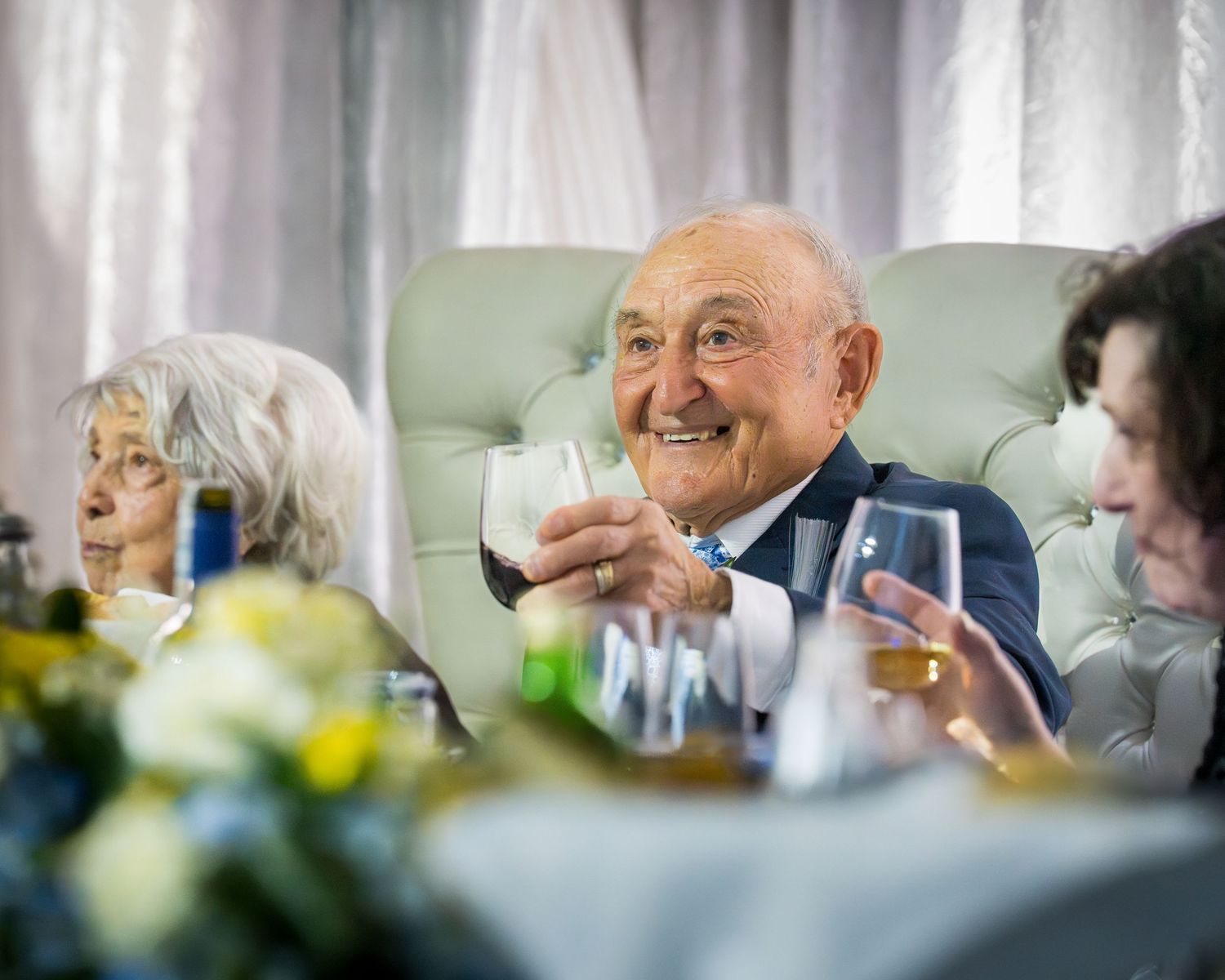 Elderly man smiling and raising a glass of red wine at a formal banquet celebration with floral centerpieces.