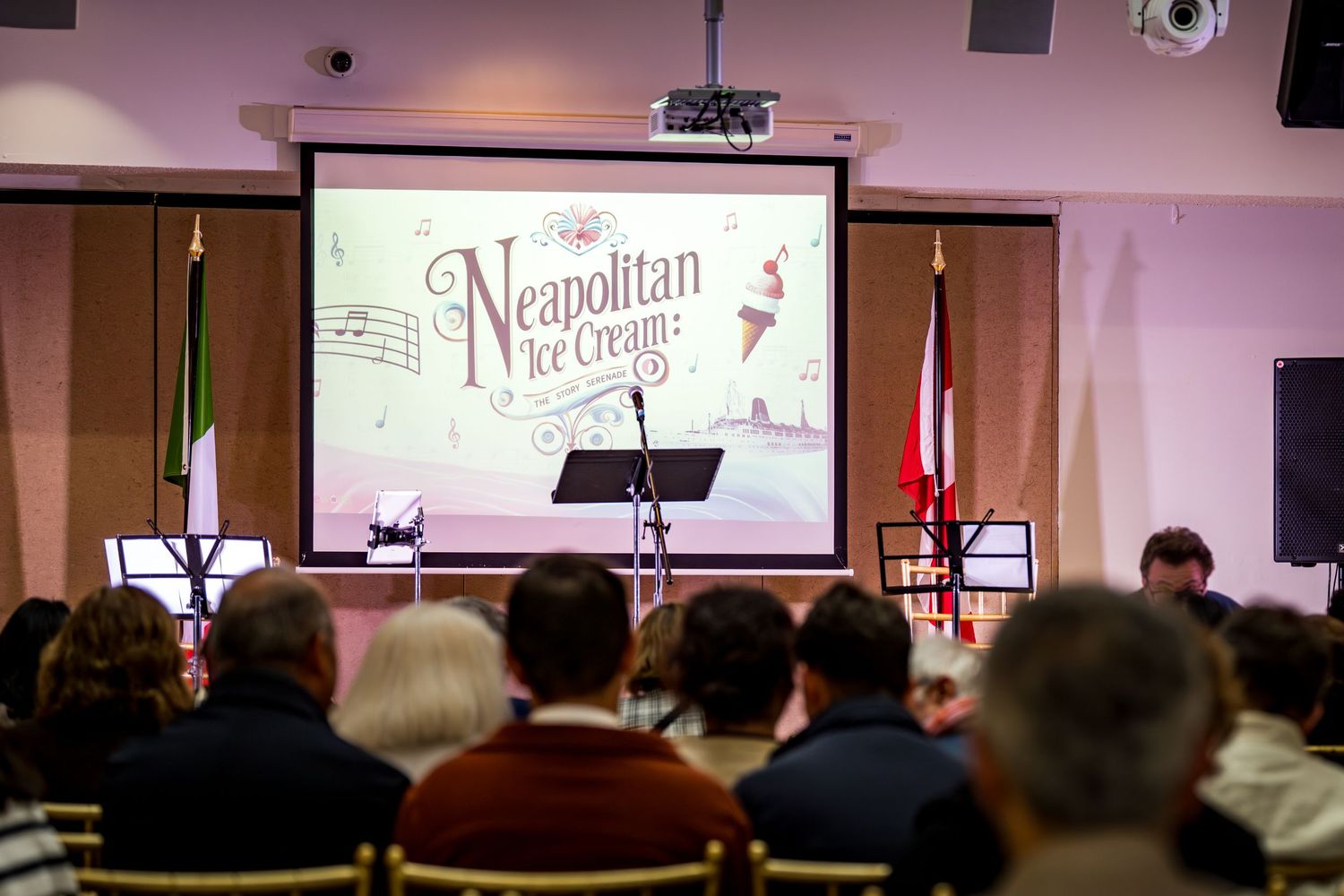Audience seated at Neapolitan Ice Cream event with presentation screen, music stands, and flags in decorated hall.