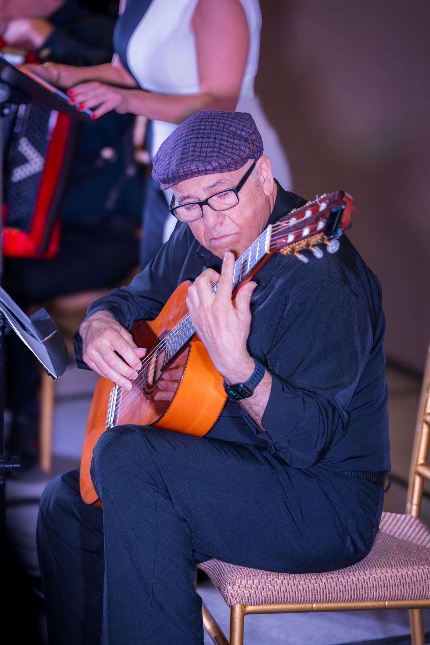 Older male guitarist wearing cap and glasses plays classical acoustic guitar on stage at live event.