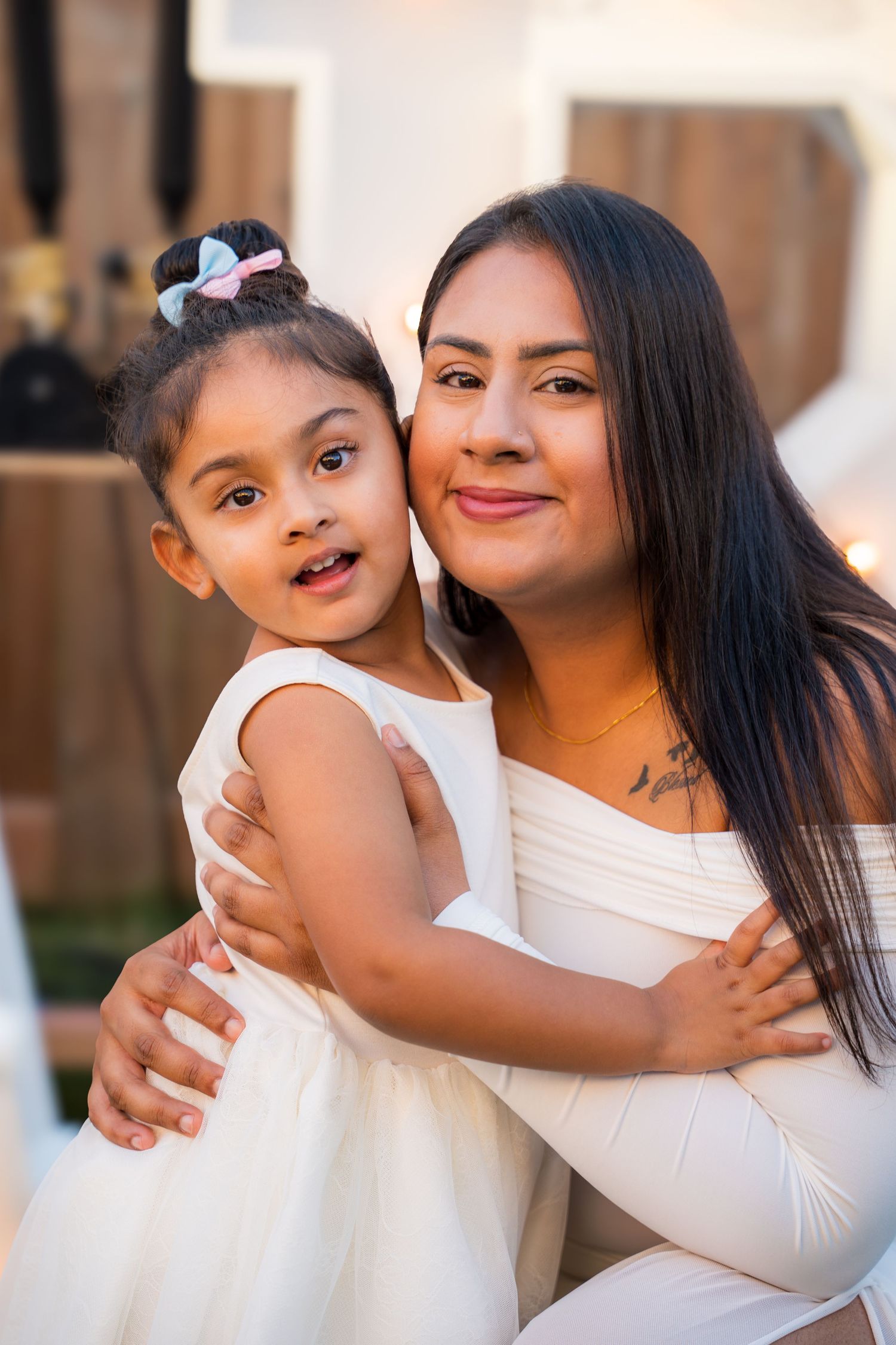 A woman in white holds a young girl in a white dress at an outdoor celebration.