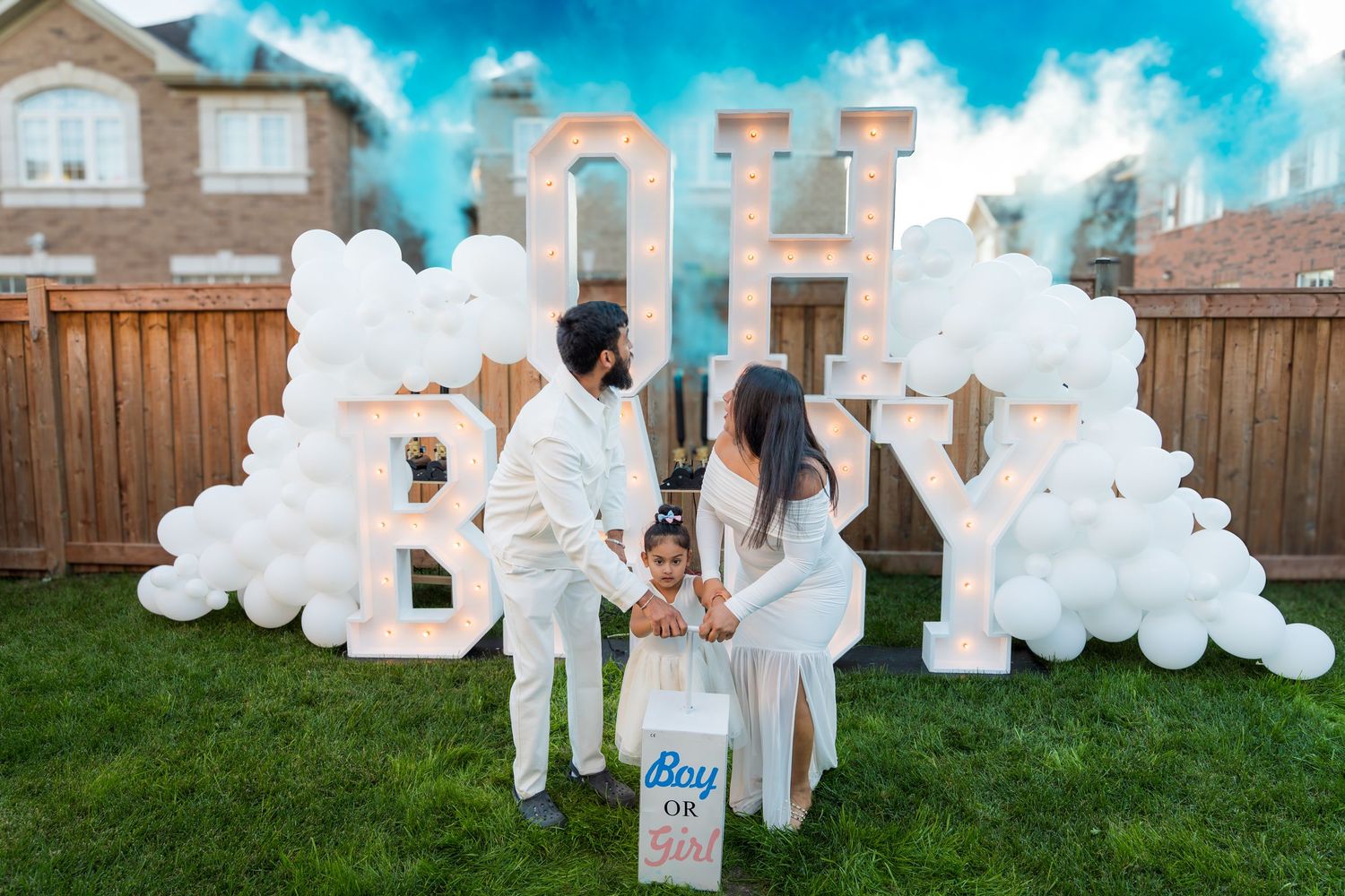 Couple with child at gender reveal party with blue smoke and OH BABY balloon backdrop.