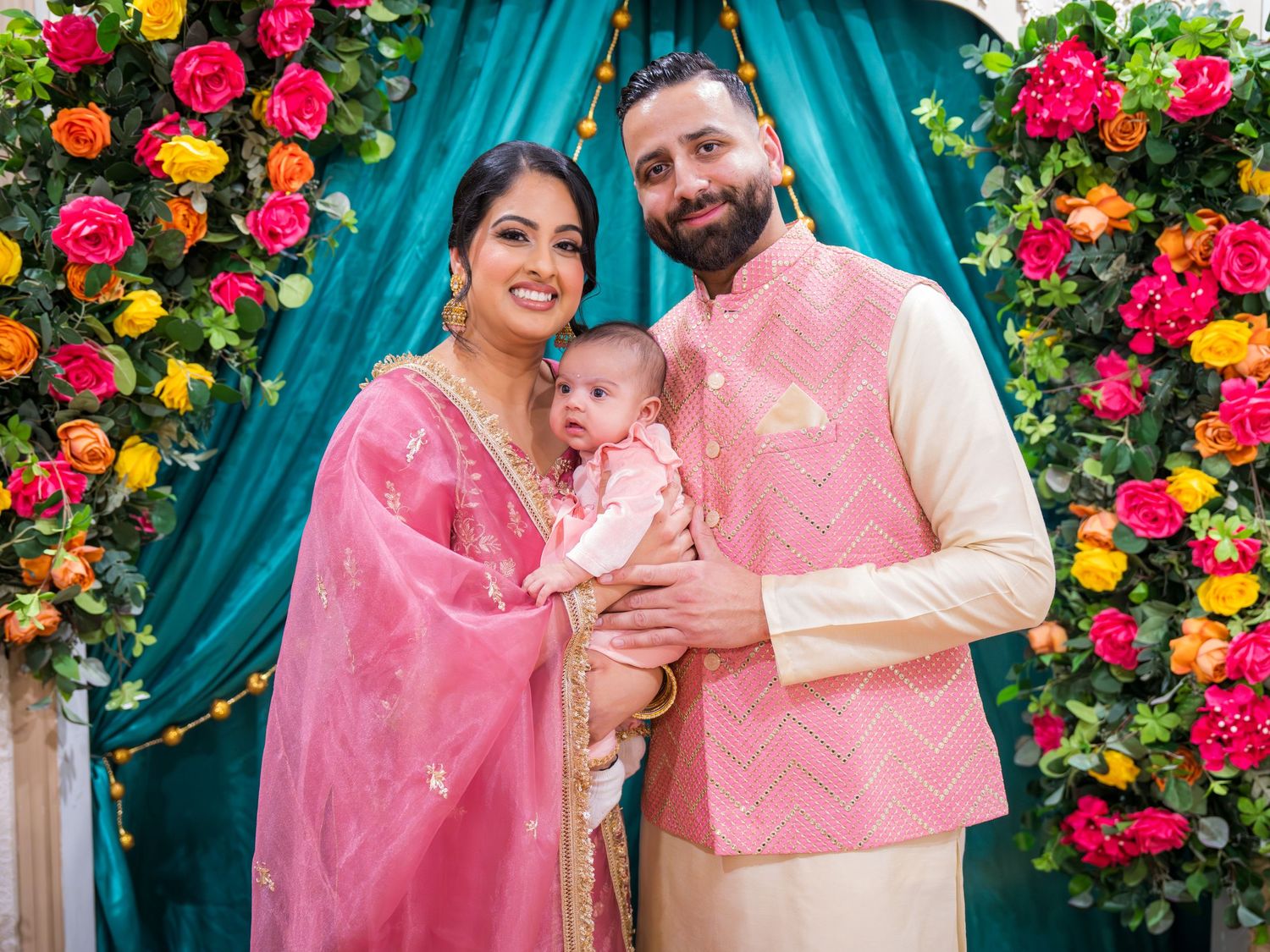 South Asian family in matching pink traditional outfits posing with newborn baby at floral ceremony.