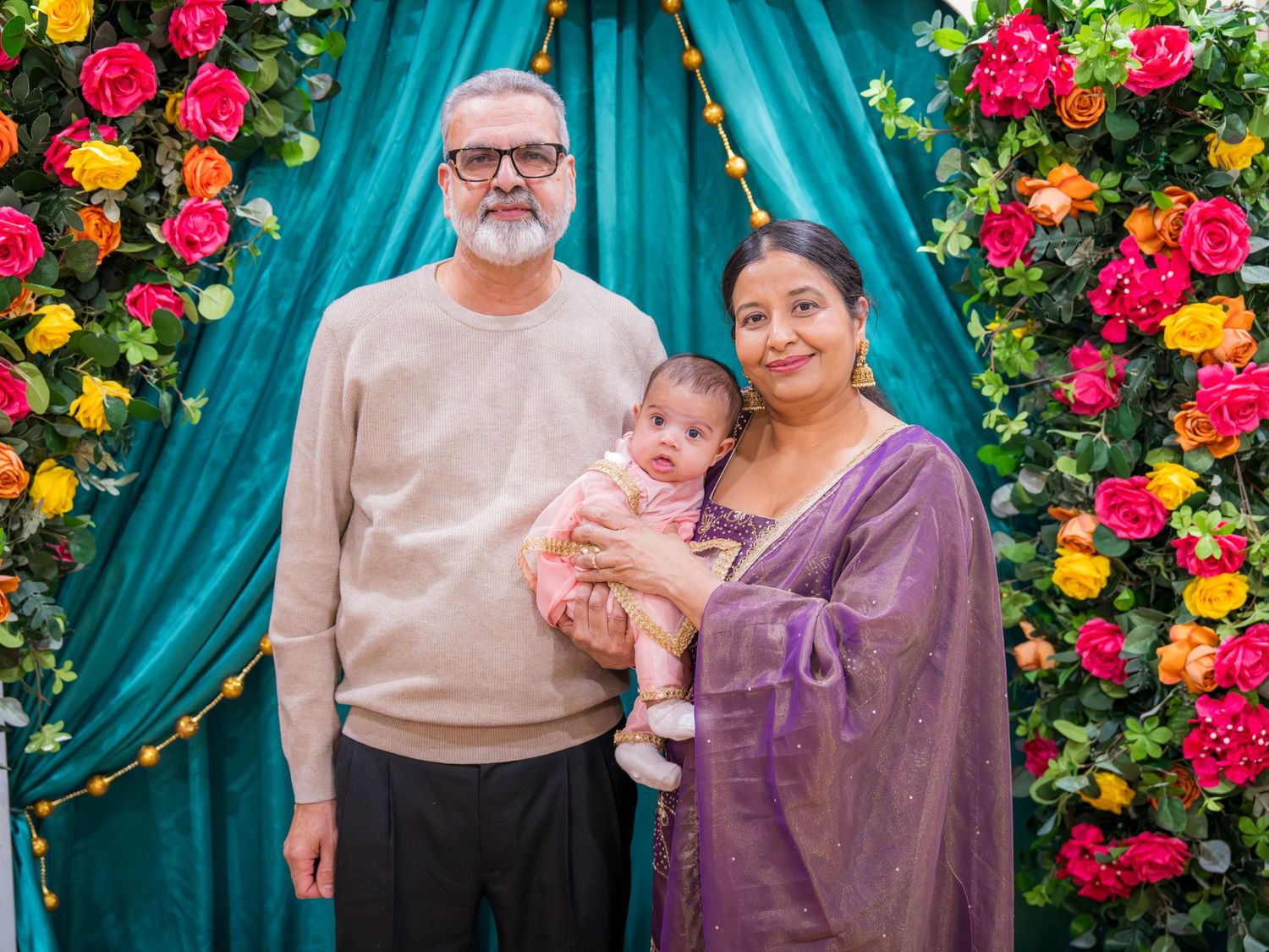 Grandparents in casual and purple saree holding newborn baby at colorful floral decorated Indian ceremony backdrop.