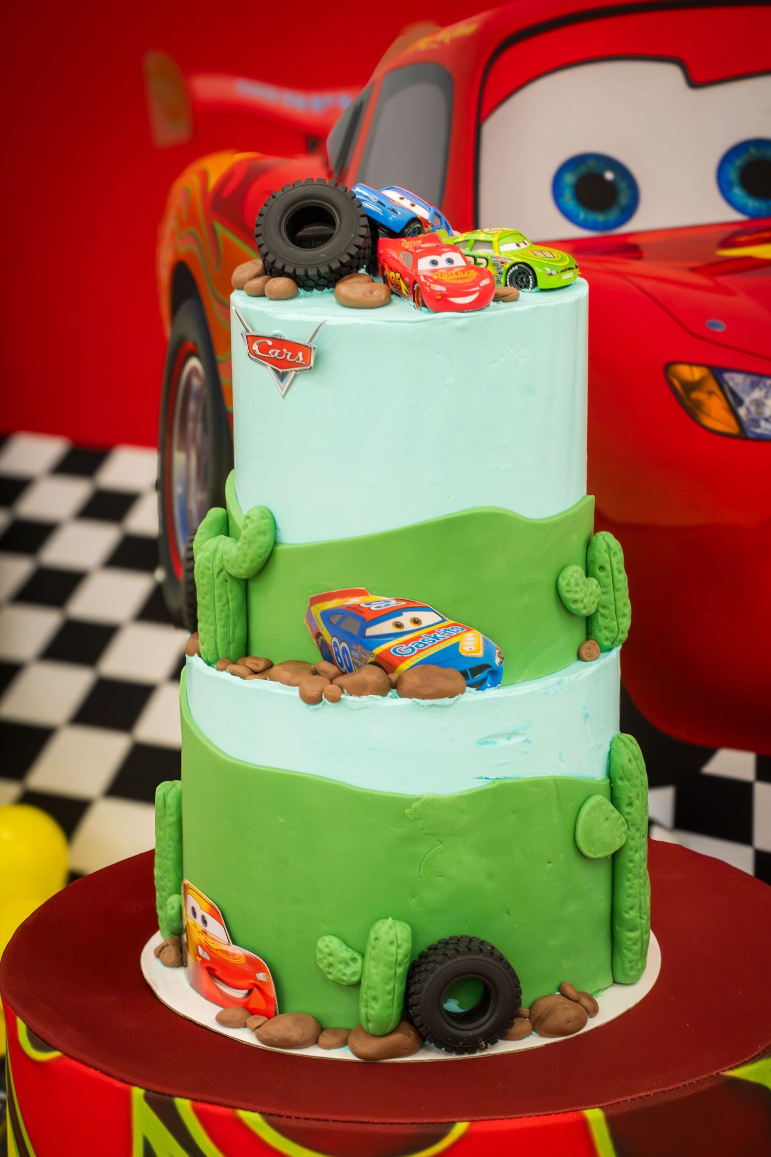 Two-tier Cars movie birthday cake featuring Lightning McQueen characters, green cacti decorations, and toy cars with red backdrop.