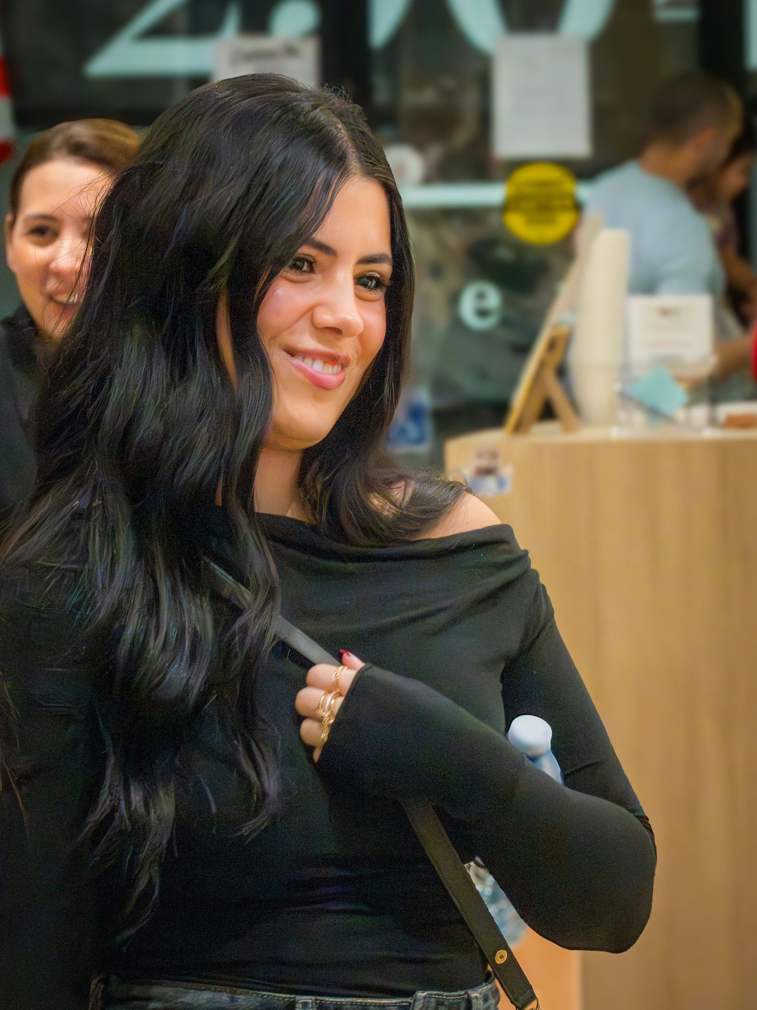 A woman with long dark wavy hair in a black off-shoulder top smiles while holding a bag strap.