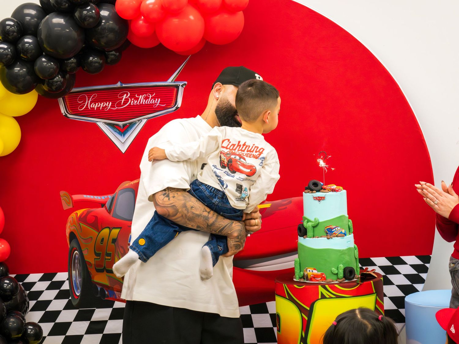 Man holding toddler near Lightning McQueen Cars themed birthday cake with sparkler candle.