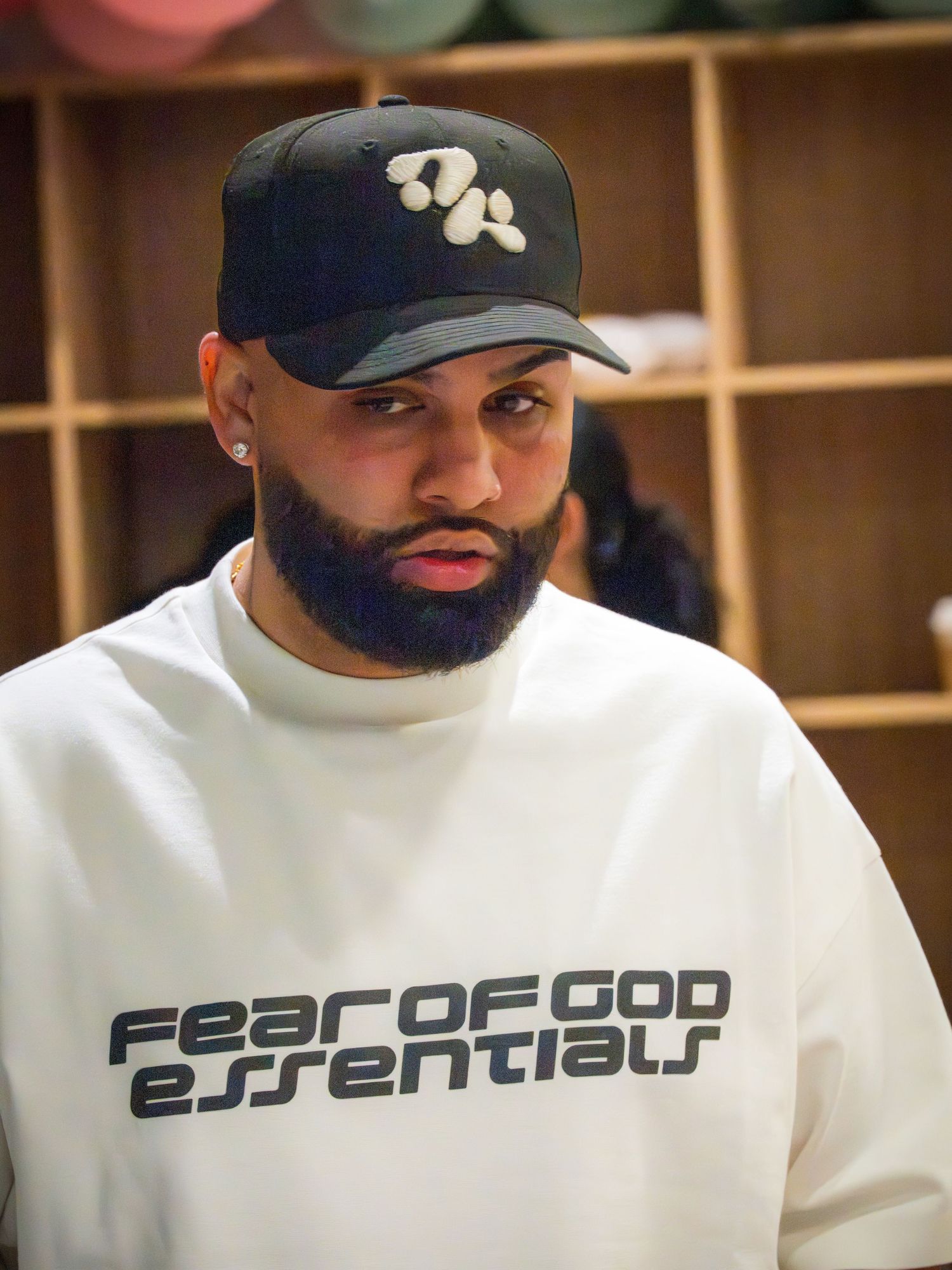 Man with beard wearing Fear of God Essentials white sweatshirt and black cap in a locker room setting.