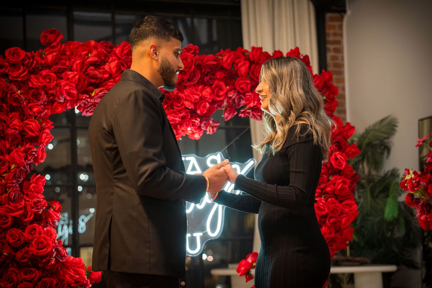 Intimate proposal setting with red rose wall and glowing neon signage in the background.