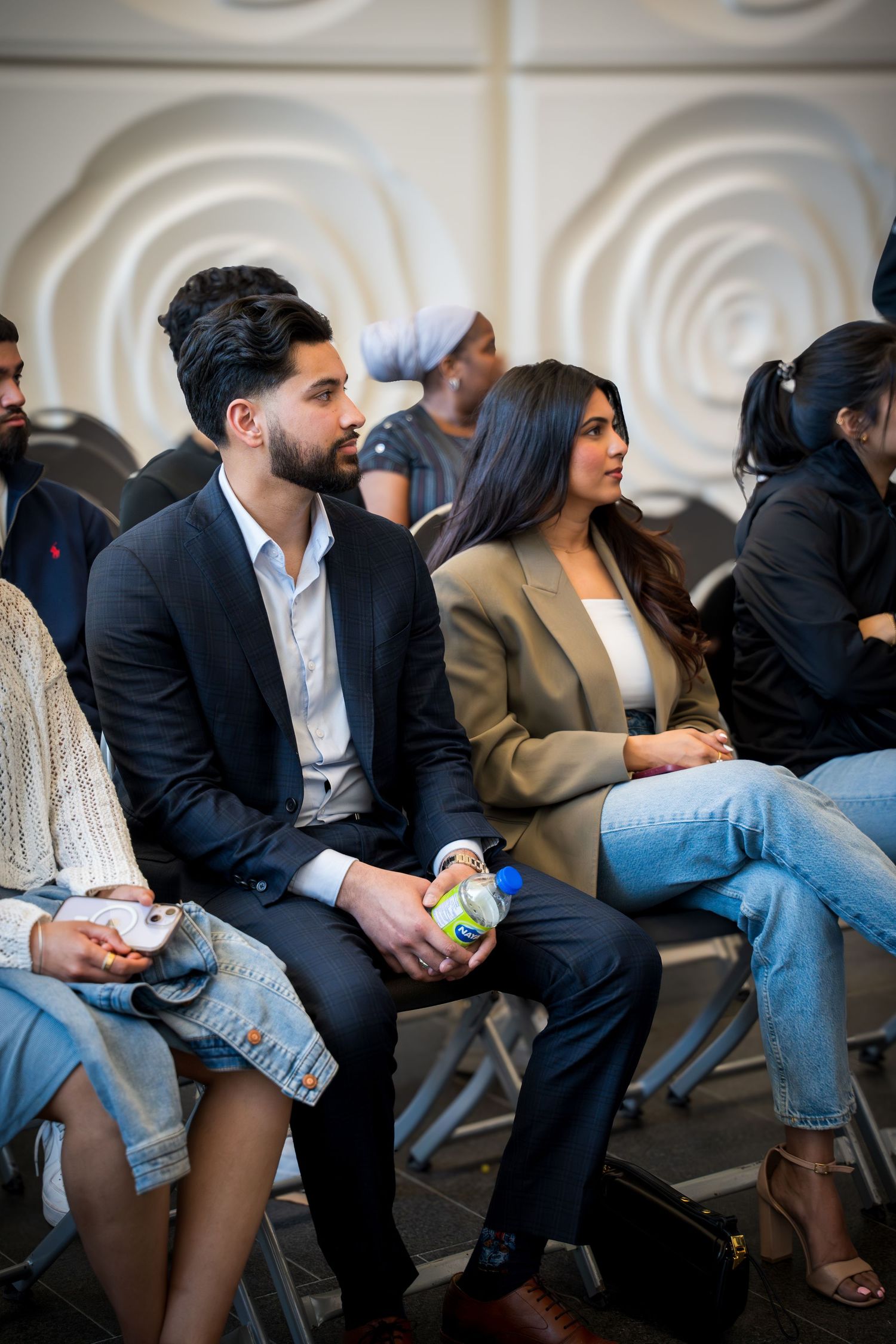 Attendees in business attire seated during presentation with decorative white wall background.