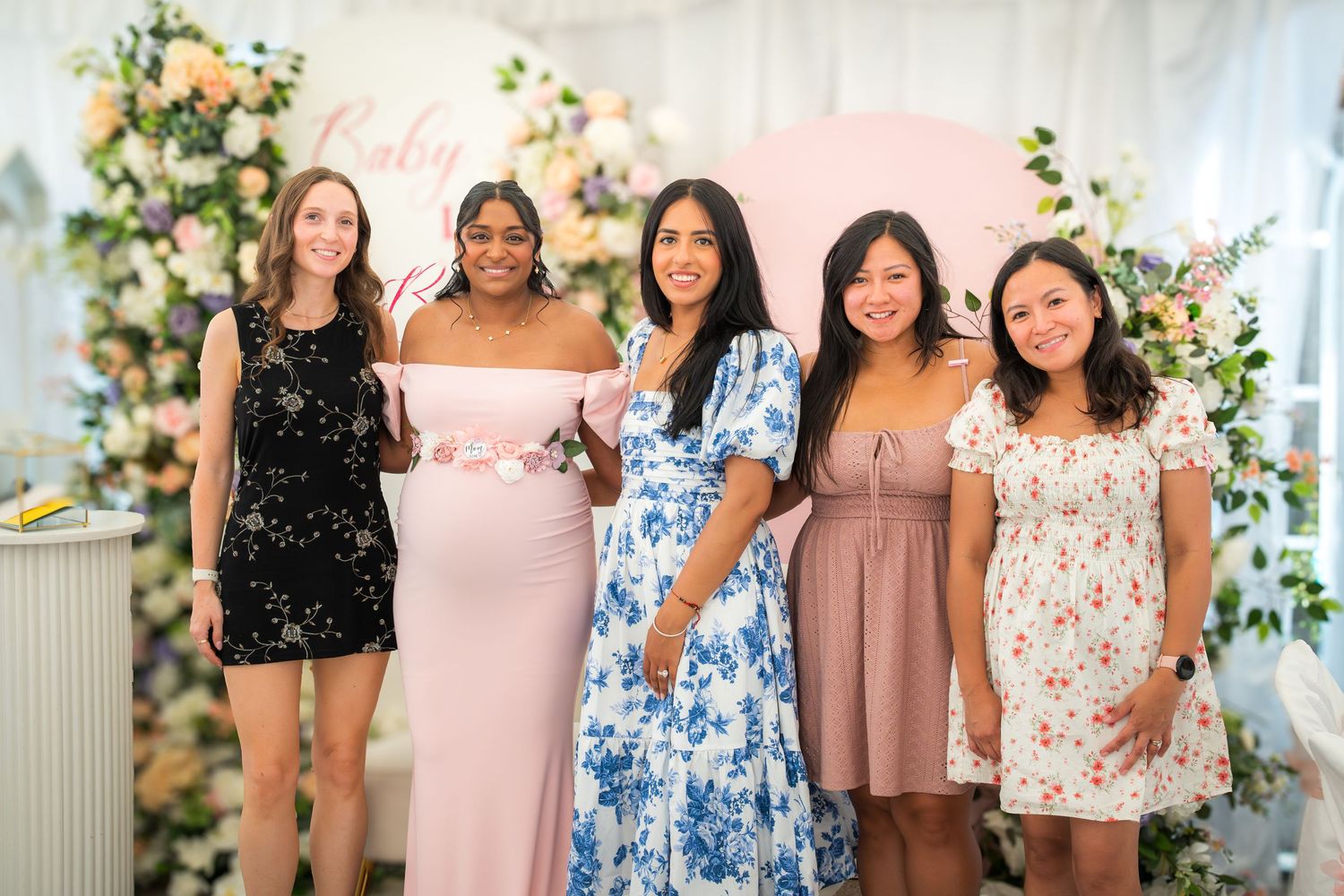Group photo with guests wearing floral and pastel dresses in front of decorated backdrop.