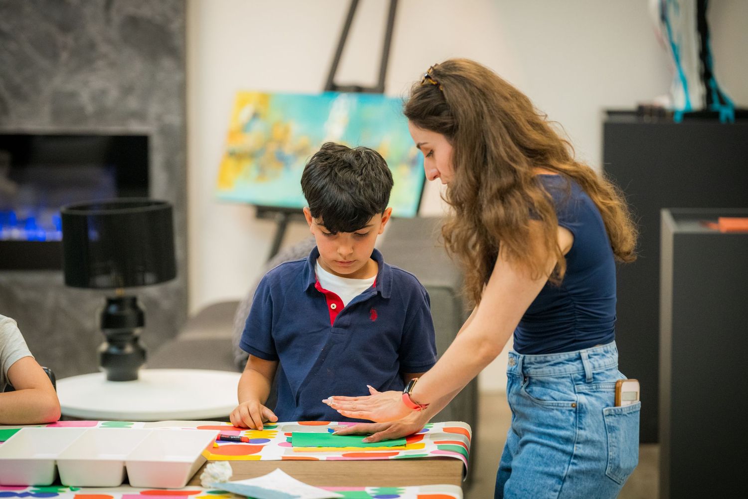 A young boy works on a colorful art project as a female teacher assists him at a table.