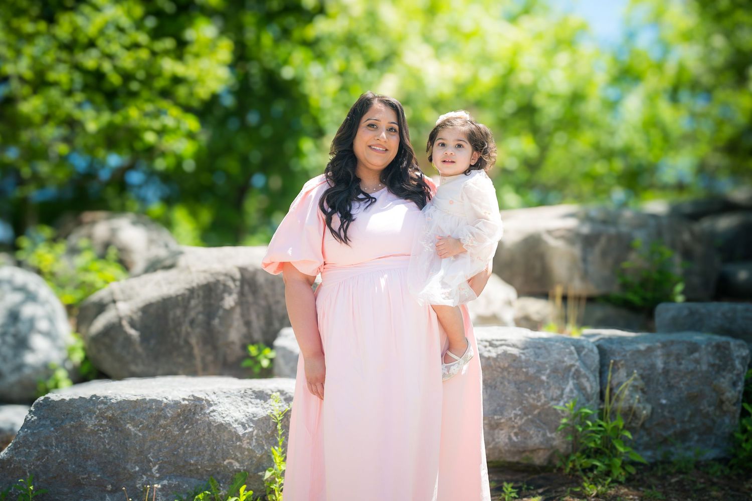 Pregnant woman in pink maxi dress holds toddler in white lace outfit outdoors among rocks and green trees.